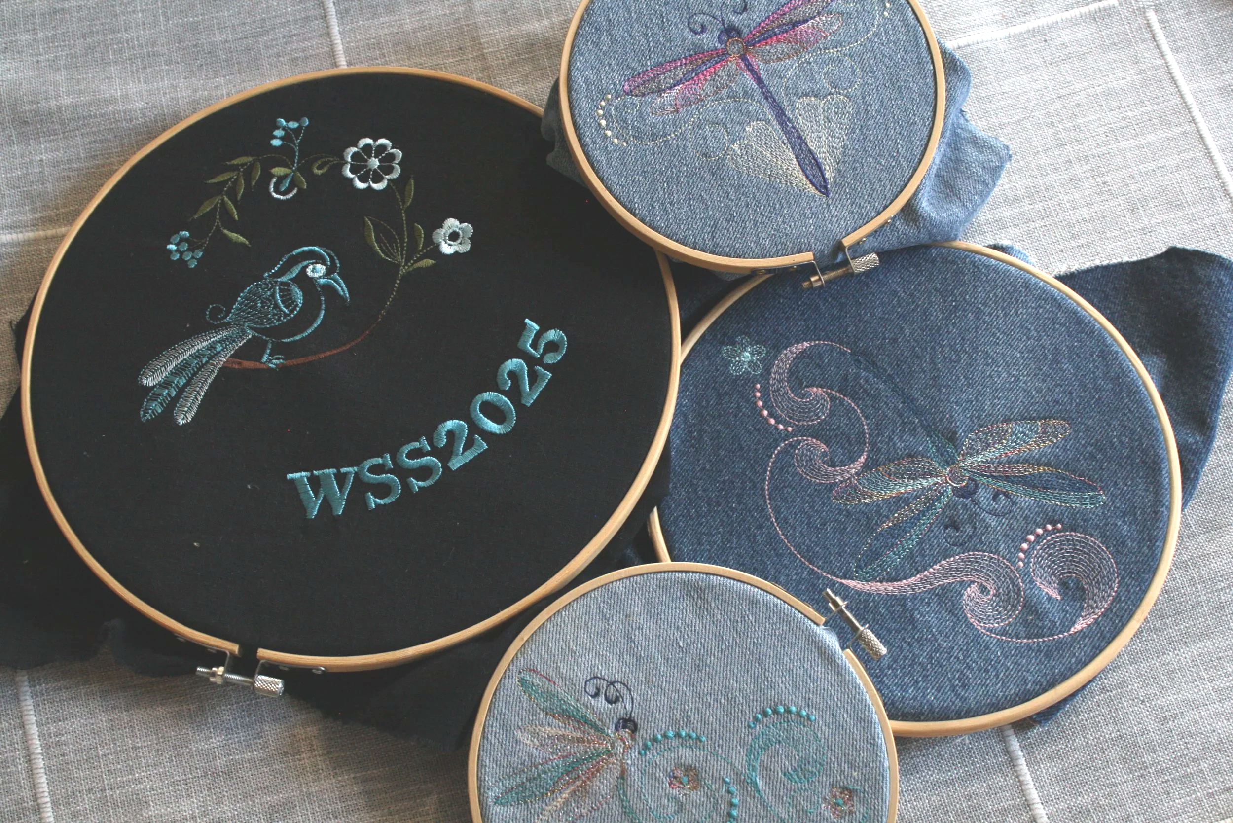 Four embroidery hoops with dragonfly and floral designs on denim and black fabric, arranged on a textured gray surface.