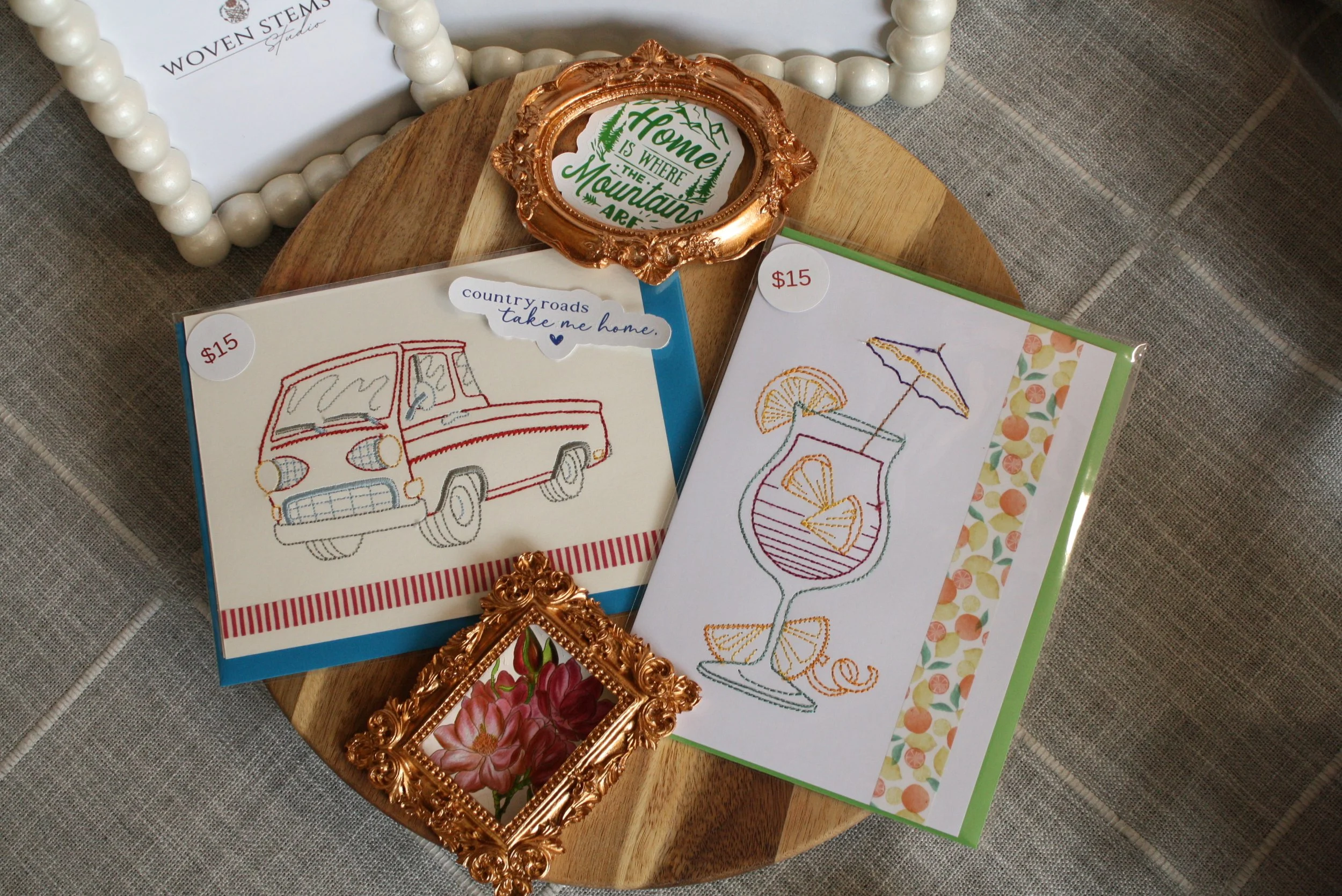 A wooden tray holding various embroidered and printed paper items, including a framed quote and greeting cards, with a decorative gold and rose gold frame and beads in the background.
