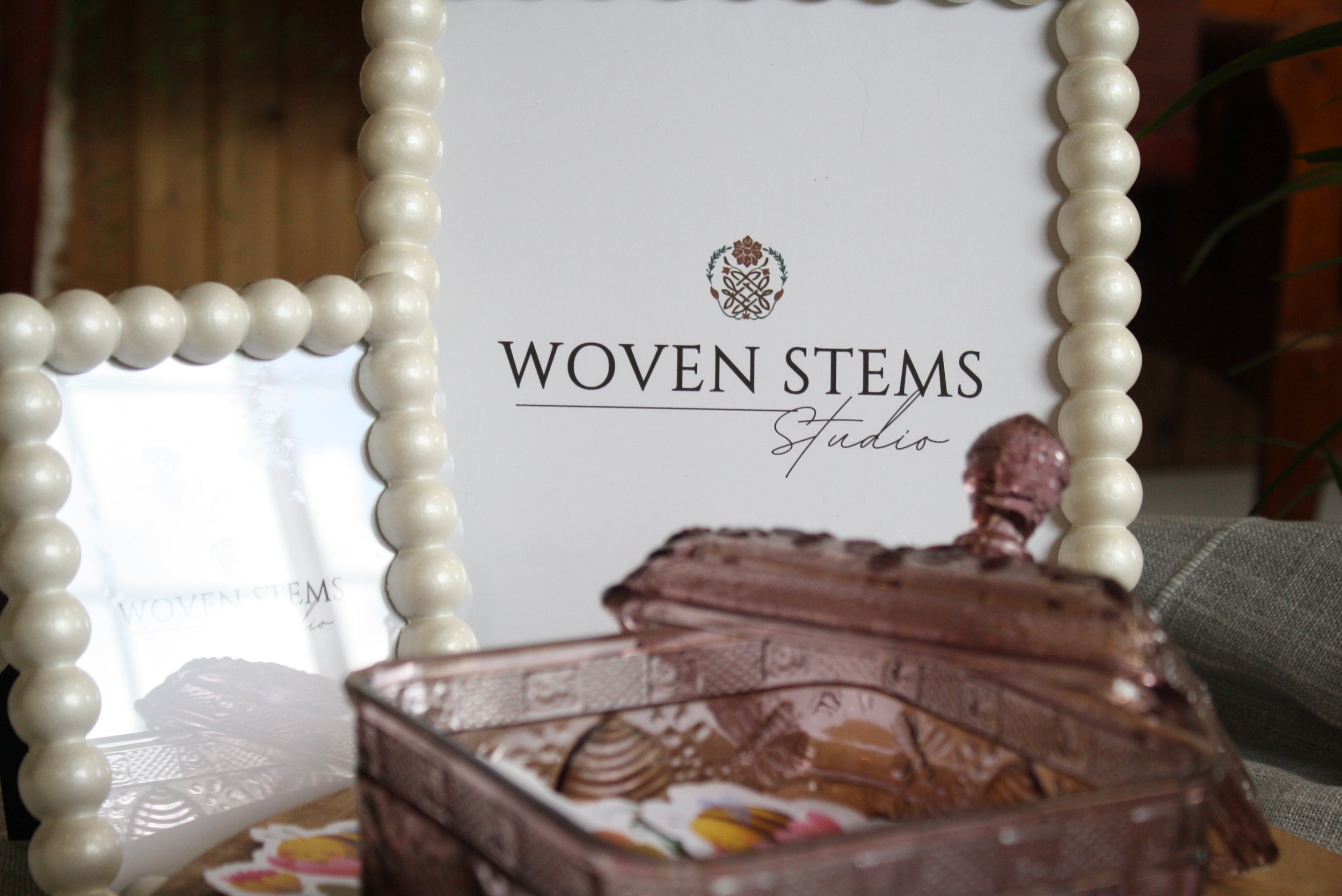 Chocolate cupcake on a plate with a swirl of frosting and a cherry on top, in front of a sign that reads "Woven Stems Studio" with pearl-like bead decorations in the background.