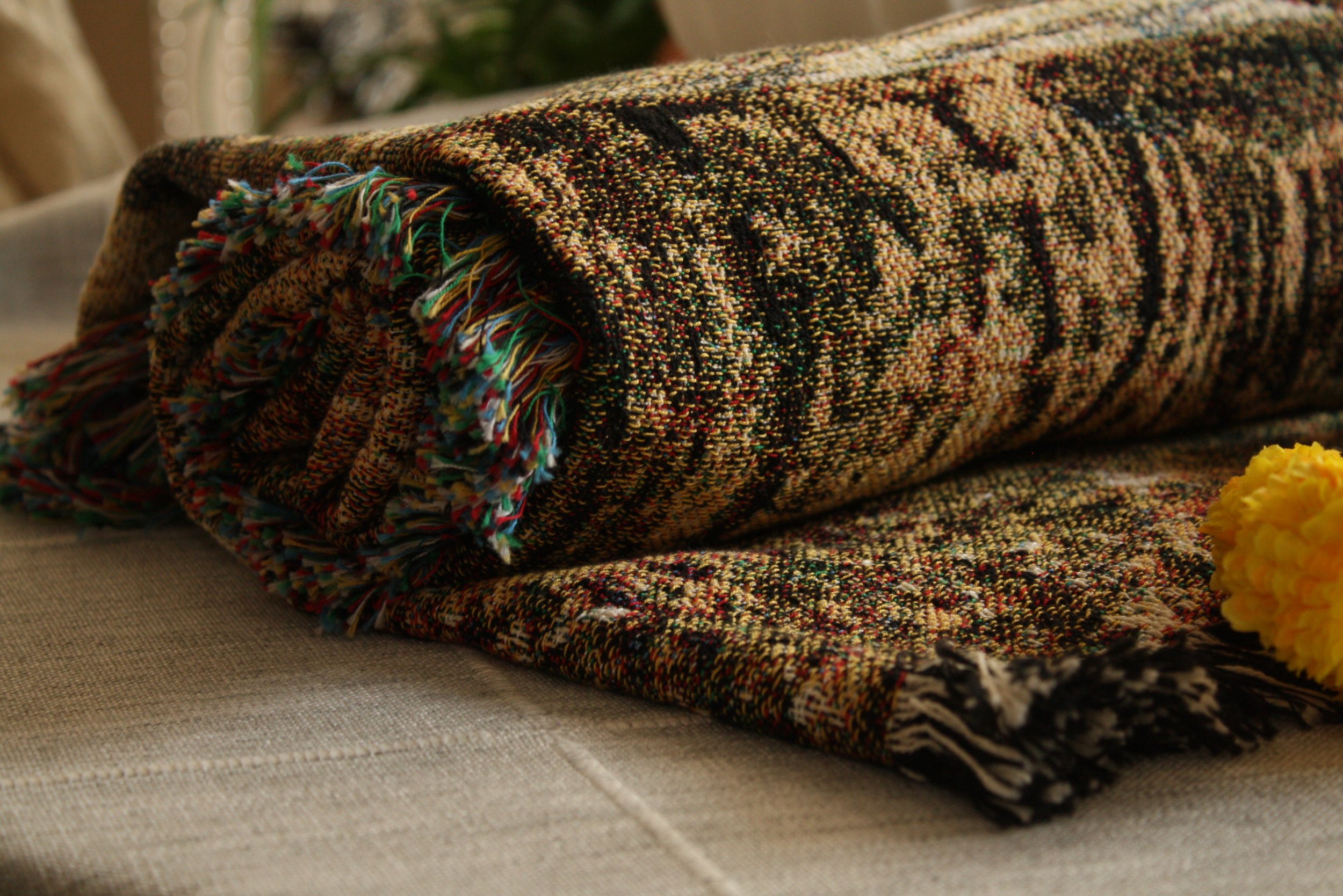 A folded multicolored woven rug with fringe edges resting on a beige fabric surface, with yellow flowers nearby.