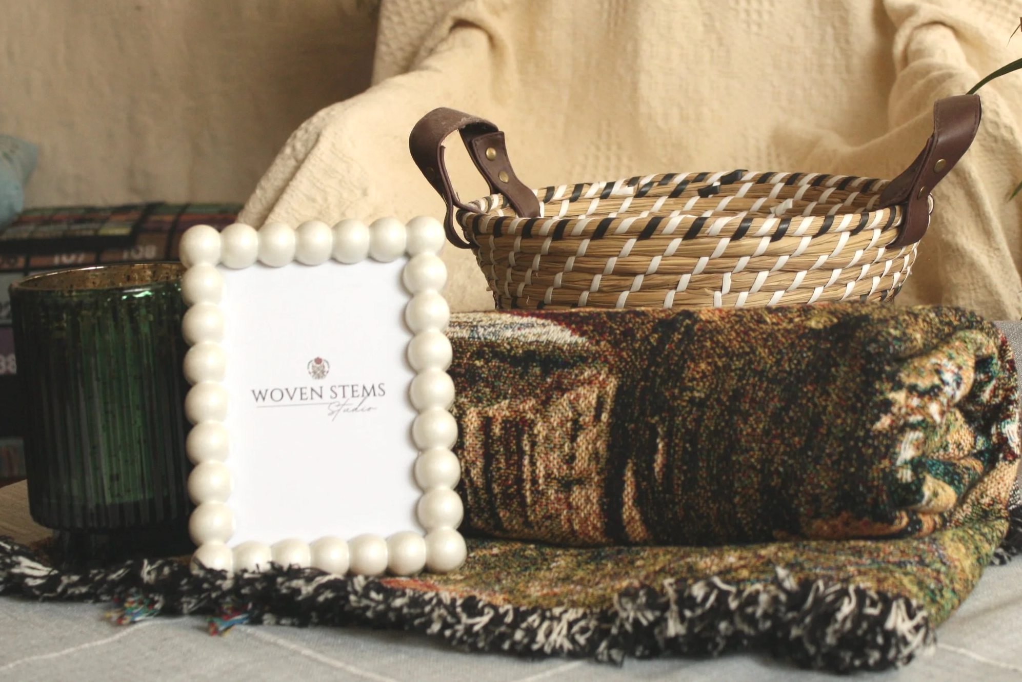 A display of various items on a colorful patterned and textured tablecloth, including a basket with leather handles, a necklace or chain with large white beads, a dark green candle holder, and a small card with partial visible text 'WOVEN STEMS' and 