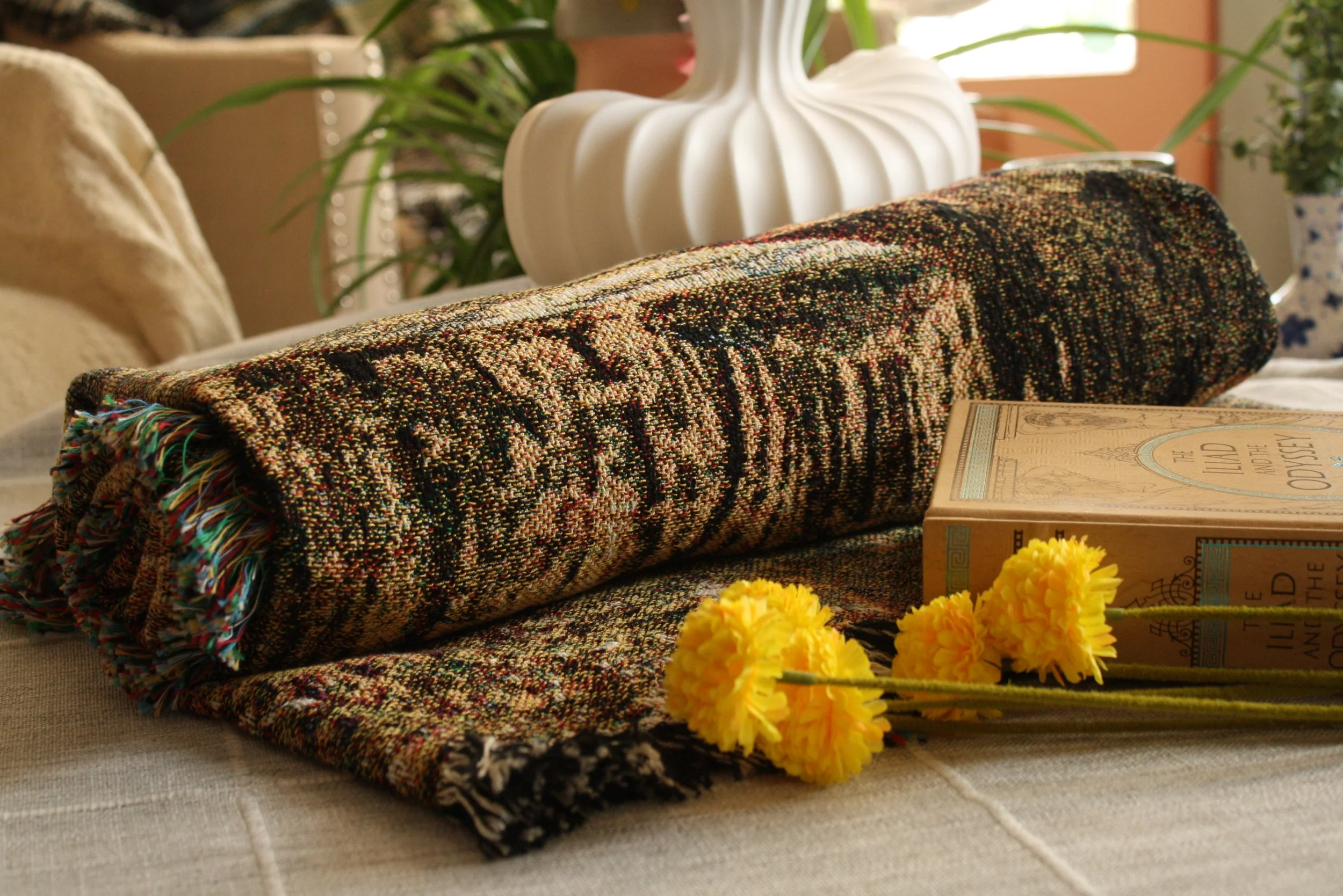 Close-up of a colorful woven rug, a book titled 'The World' is partially visible, and yellow flowers in foreground, with plants and a white decorative vase in the background.