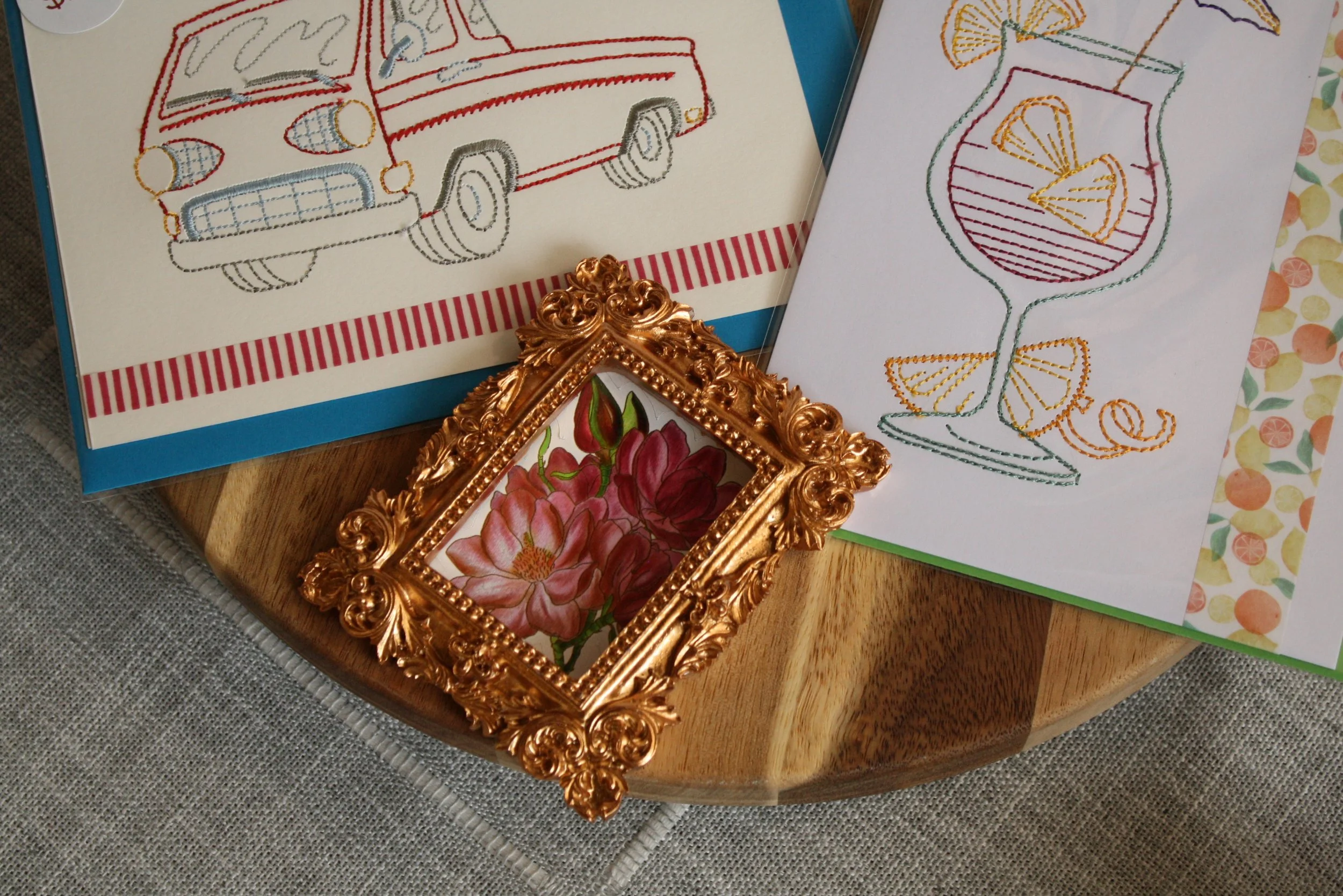 Colorful embroidered cards with summer and travel themes, including a car, a beach ball, and a cocktail glass, displayed on a wooden surface with a decorative gold picture frame.