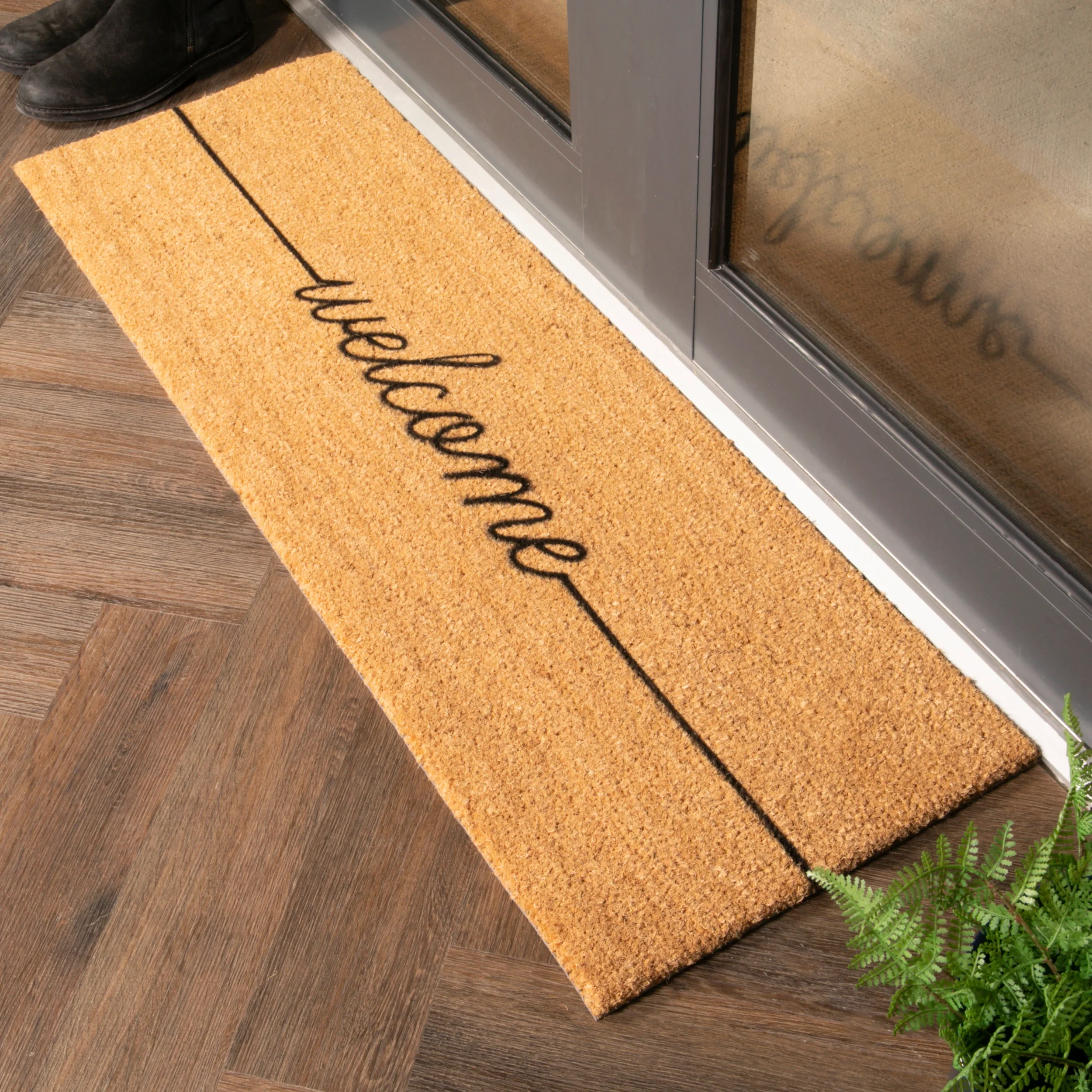 A door mat with the word "welcome" written in cursive, placed on a wooden floor near a glass door.
