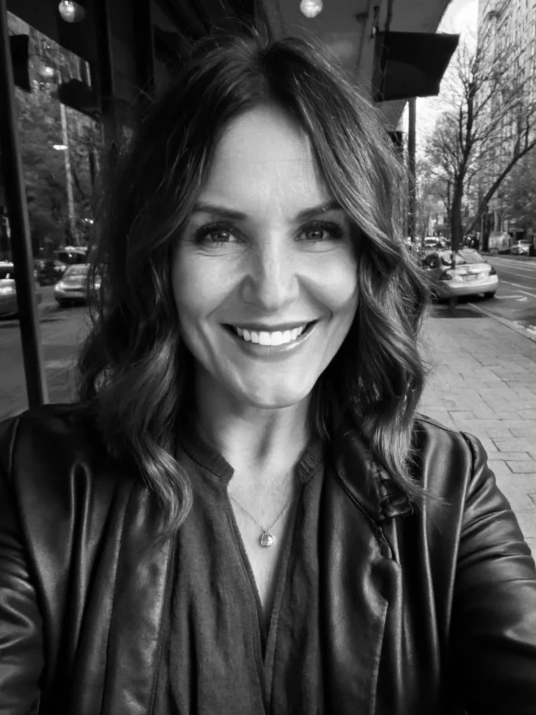 A woman with wavy shoulder-length hair smiling for a selfie outside a storefront. She is wearing a leather jacket and a necklace with a small pendant, with cars and trees in the background.