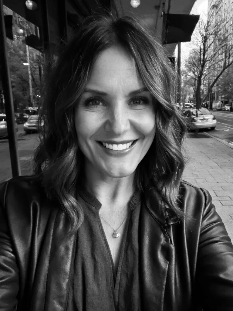 A smiling woman with wavy hair taking a selfie outside on a sidewalk with parked cars and trees in the background in black and white.