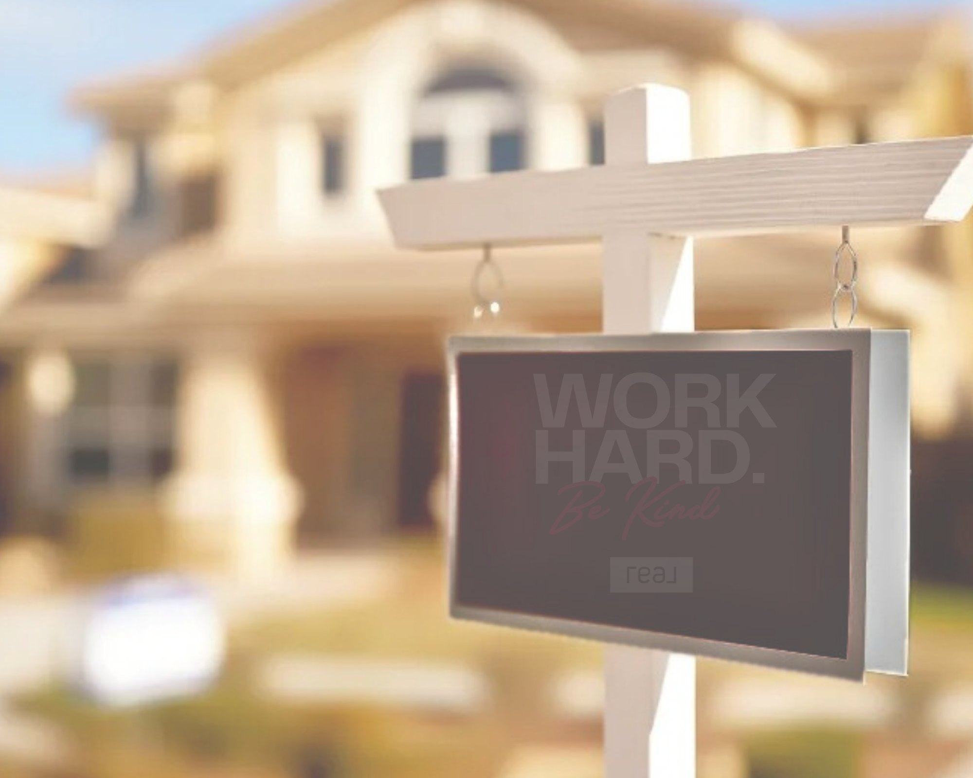 A small blackboard sign hangs on a white post in front of a large house, with the words "Work Hard. Be Kind" written on it. The house in the background appears to be a modern beige-colored building with multiple windows.