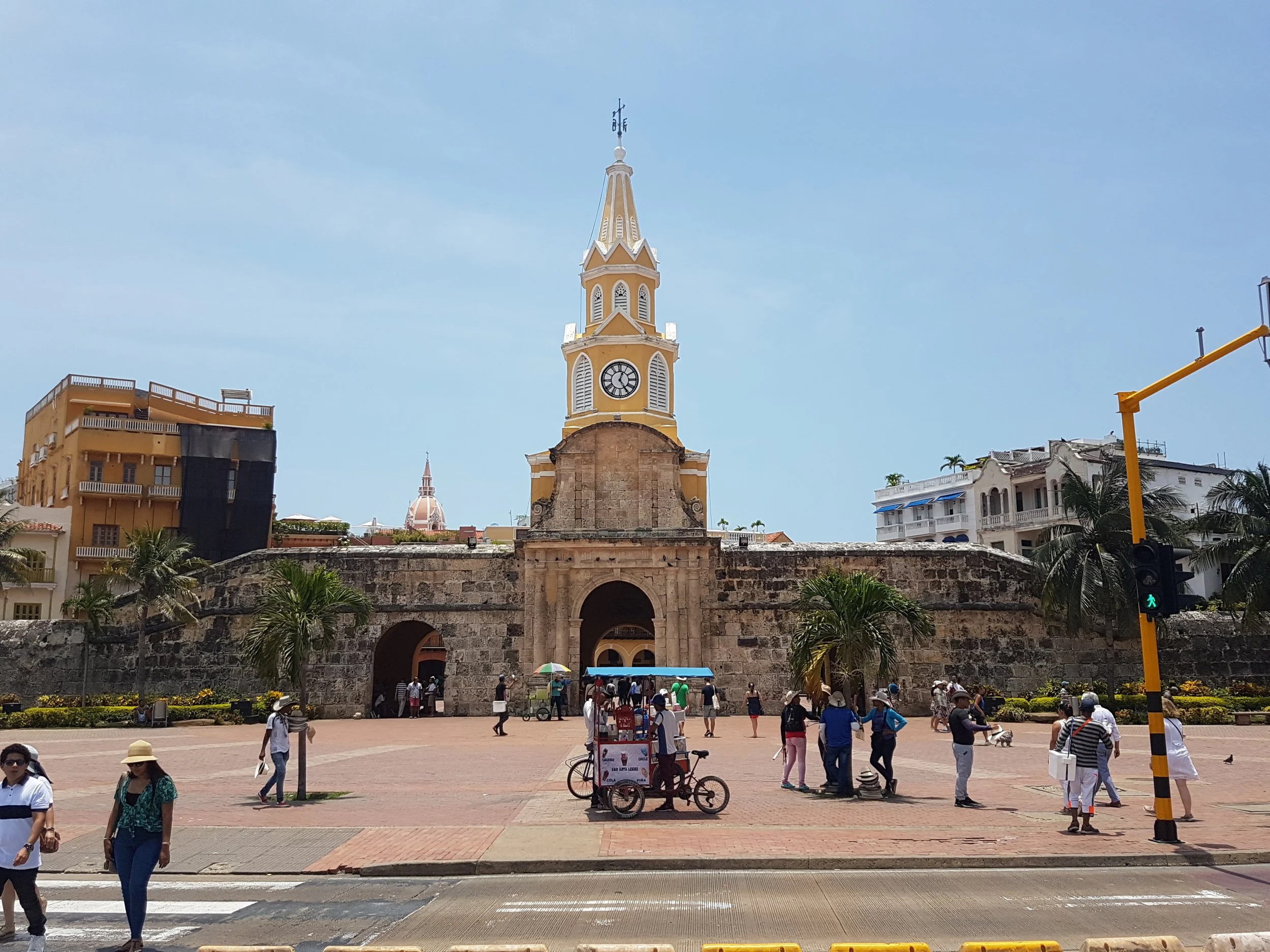 Historical clock tower with yellow and white exterior, arched entrance, and a tall spire in a busy city square with people walking and colorful vendors.