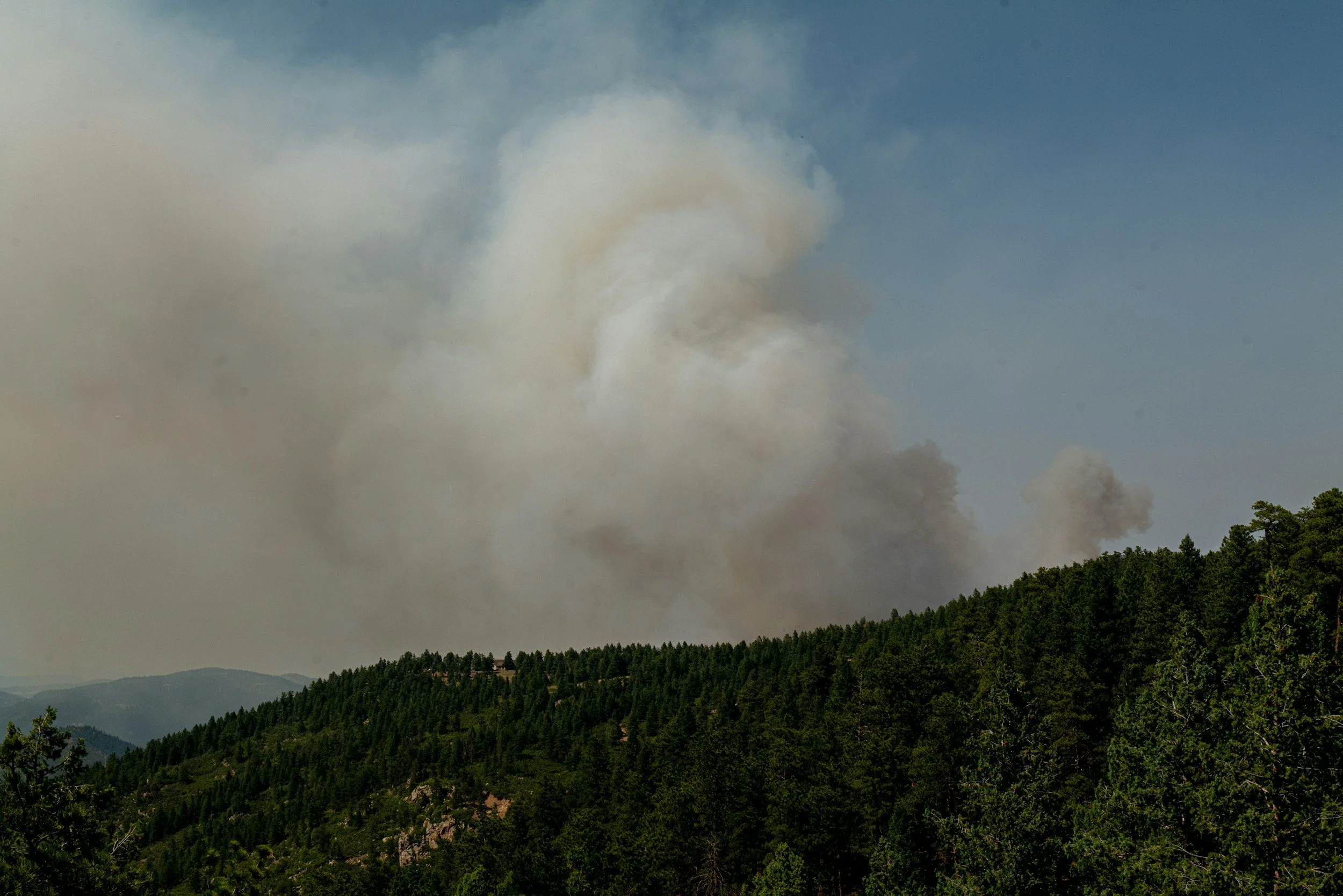 Thick gray and white smoke billowing from a wildfire in a forested mountain area under a partly cloudy sky.