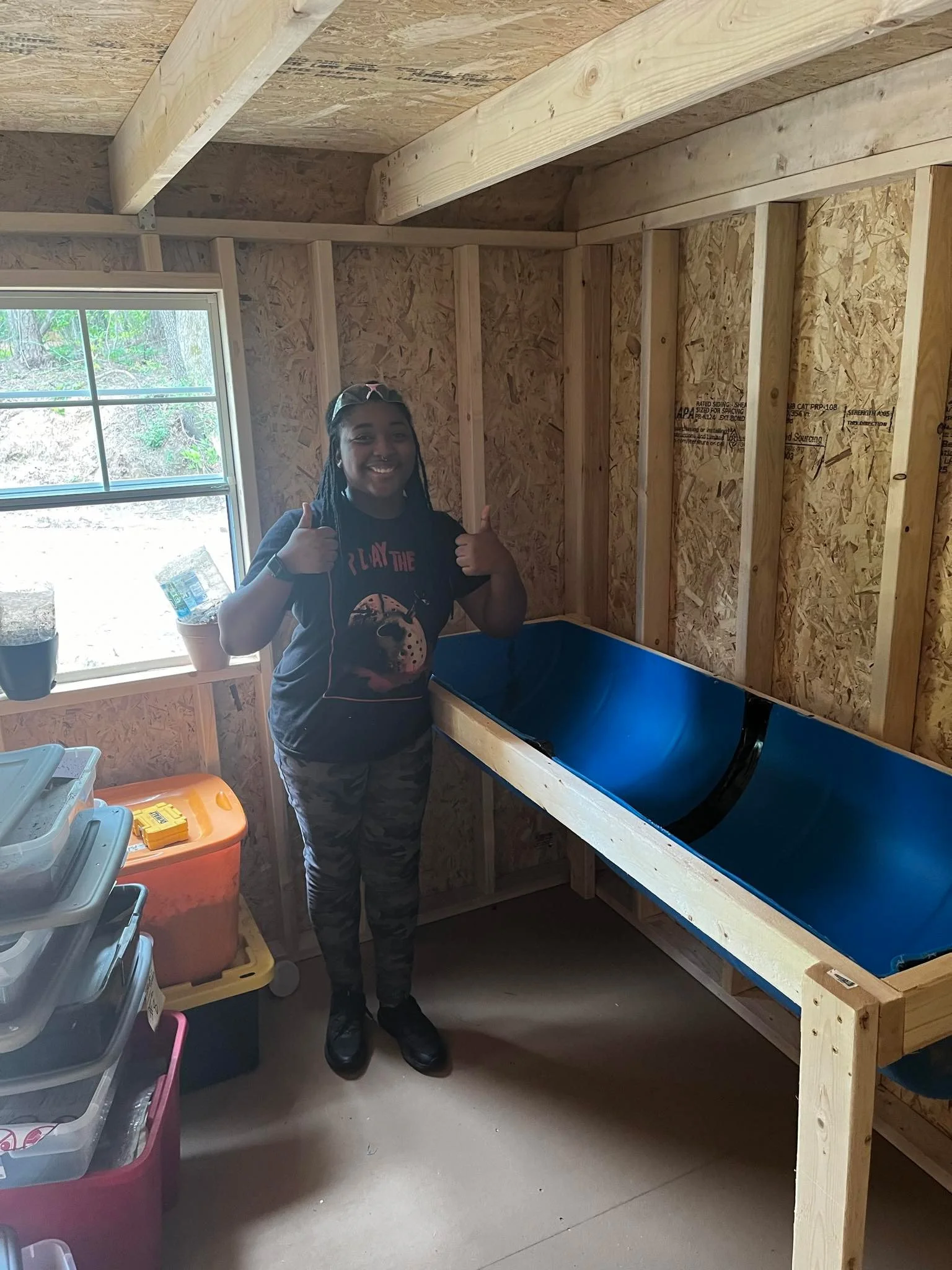 A woman standing inside a wooden shed or workshop next to a blue tub, smiling and giving a thumbs-up with both hands.