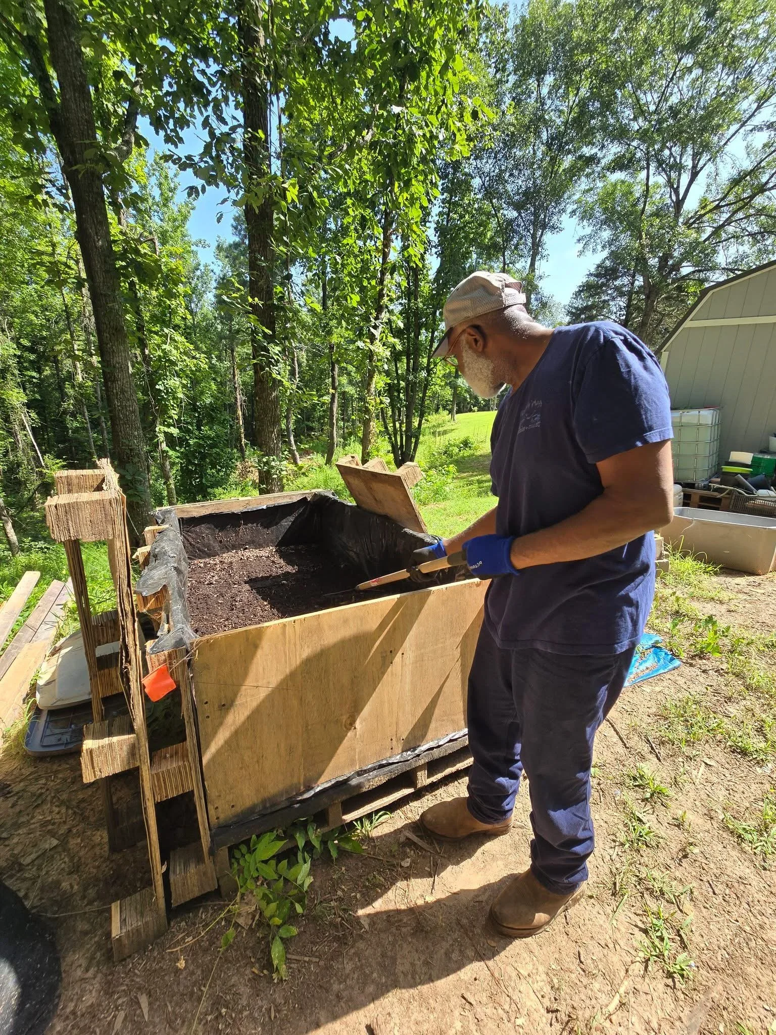 A man wearing a cap, gloves, and sunglasses is working outdoors in a wooded area, tending to a large wooden compost bin filled with soil or compost material.