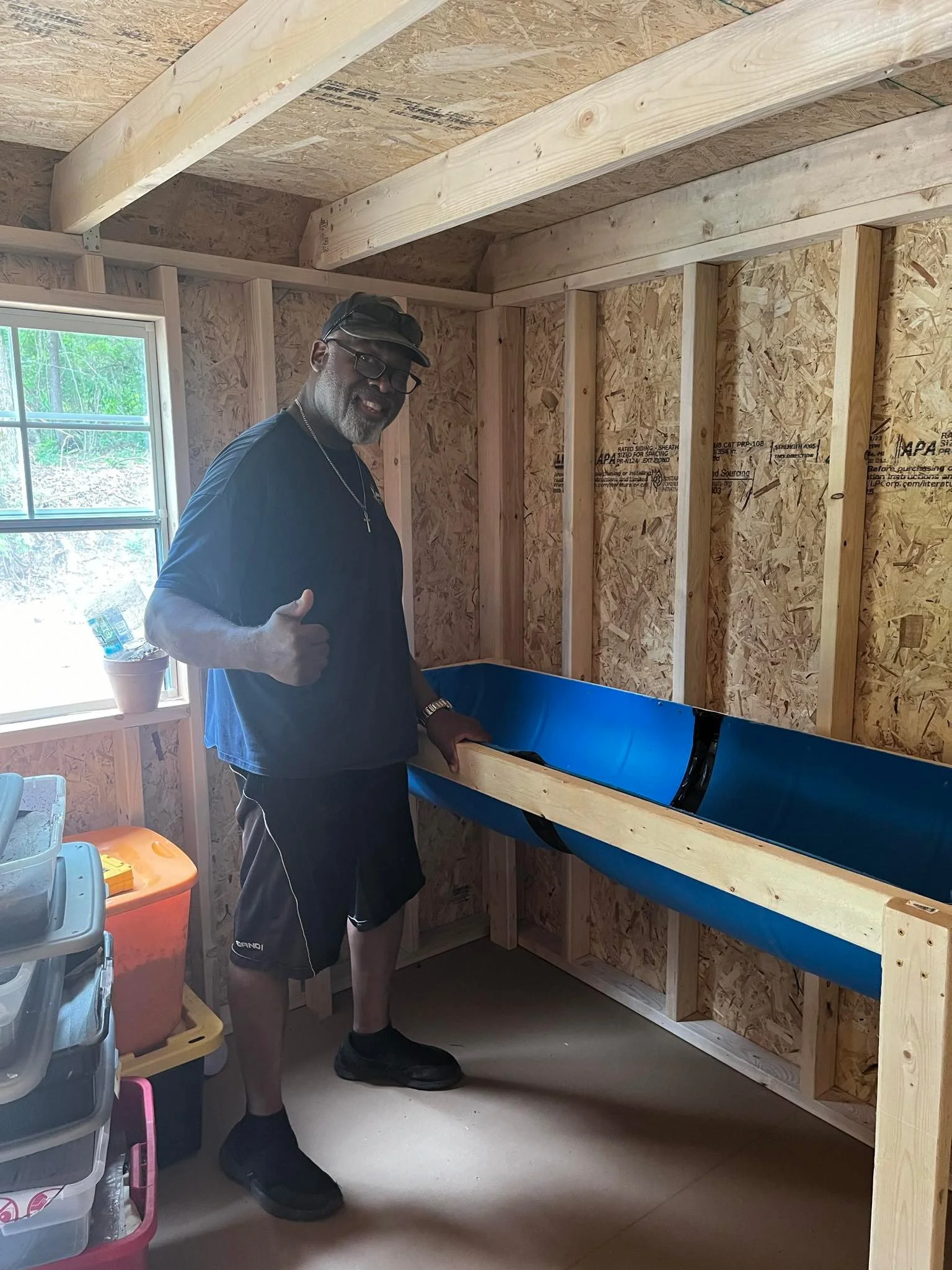 A man giving a thumbs-up inside a wooden shed or workshop, standing next to a partially assembled blue boat or kayak.