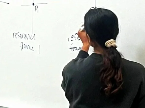 A group of children learning about renewable energy from a male teacher in a classroom, with a small plant, wind turbine models, and recycling bins in the background.
