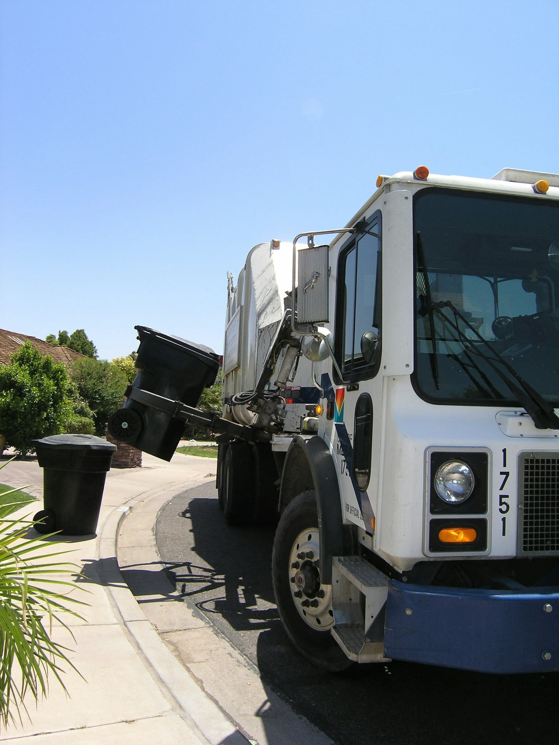 Garbage truck collecting trash from a black trash bin on a curb during daytime with a blue sky.