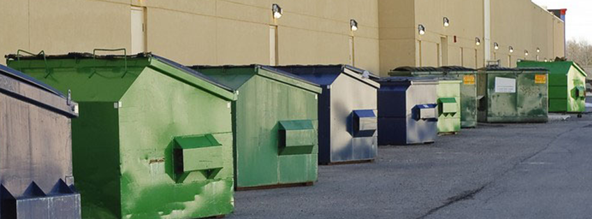 A row of large, green, and gray commercial dumpsters lined up along a paved area against a beige building wall.