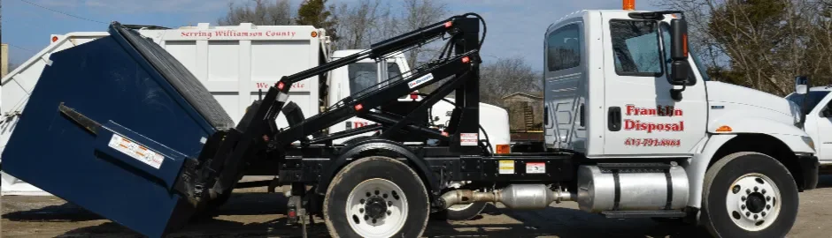 White garbage truck with black lifting arm and blue trash bin on a sunny day.