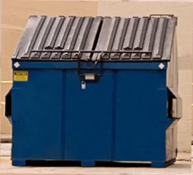A large industrial dumpster with a gray metal lid, blue body, and black handles, positioned against a beige wall.