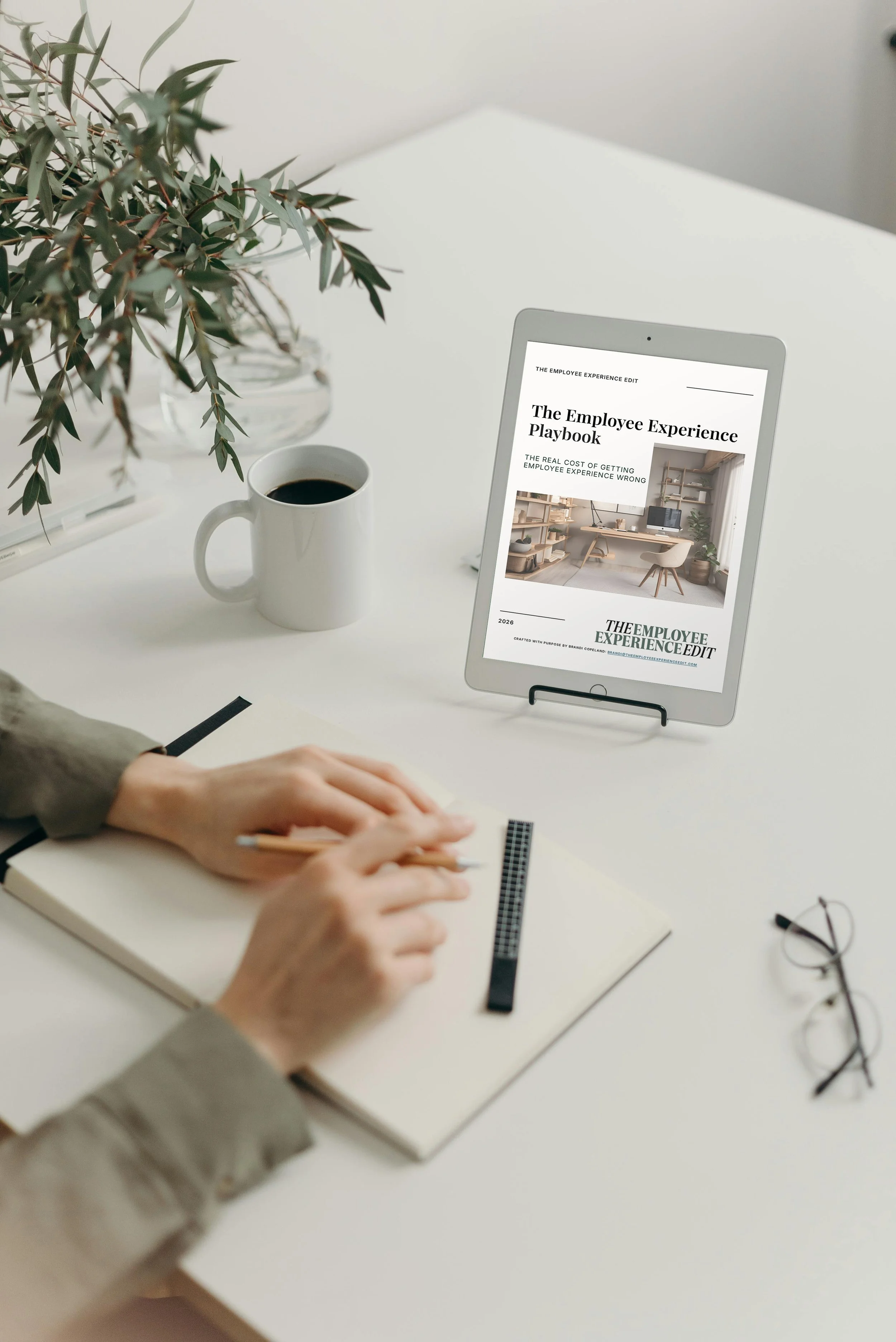 A person writing in a notebook at a white desk with a cup of coffee, a tablet displaying a report on employee experience, a pair of glasses, and a plant in a glass vase.