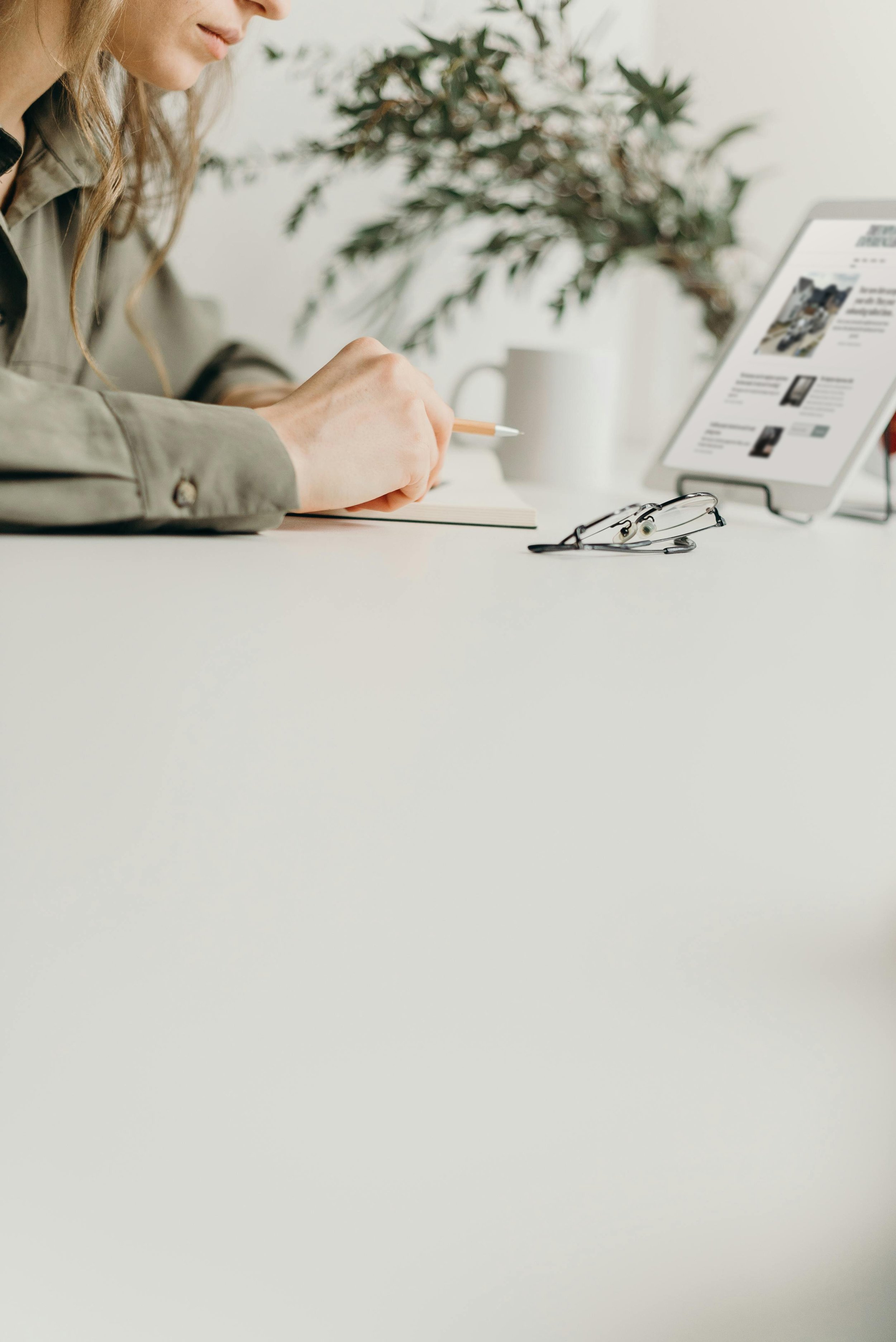 Person writing in a notebook at a desk with glasses on the table and a tablet device displaying a webpage.