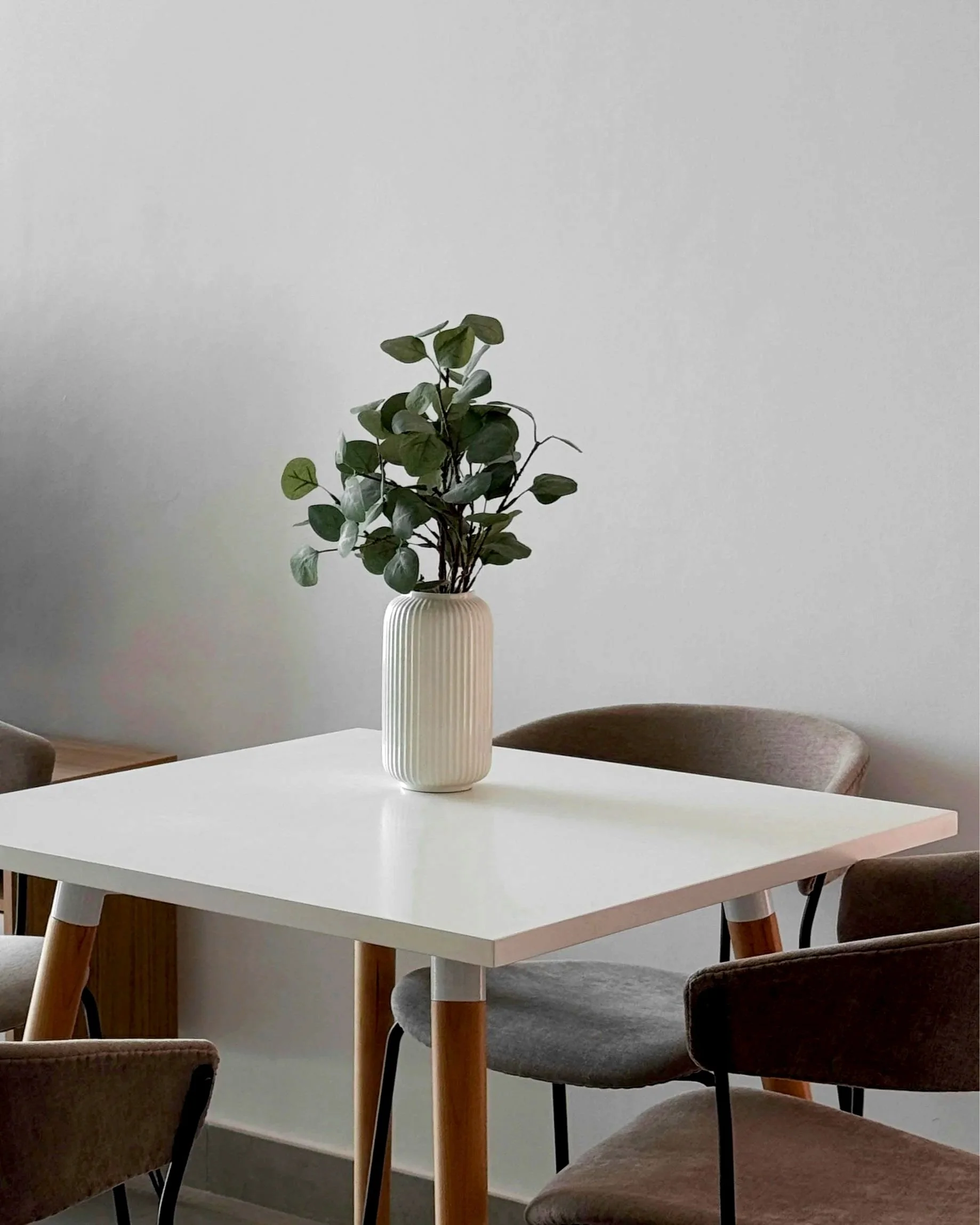 White dining table with wooden legs, centered with a white ribbed vase holding green leafy plant, surrounded by upholstered chairs in neutral tones, against a plain light gray wall.