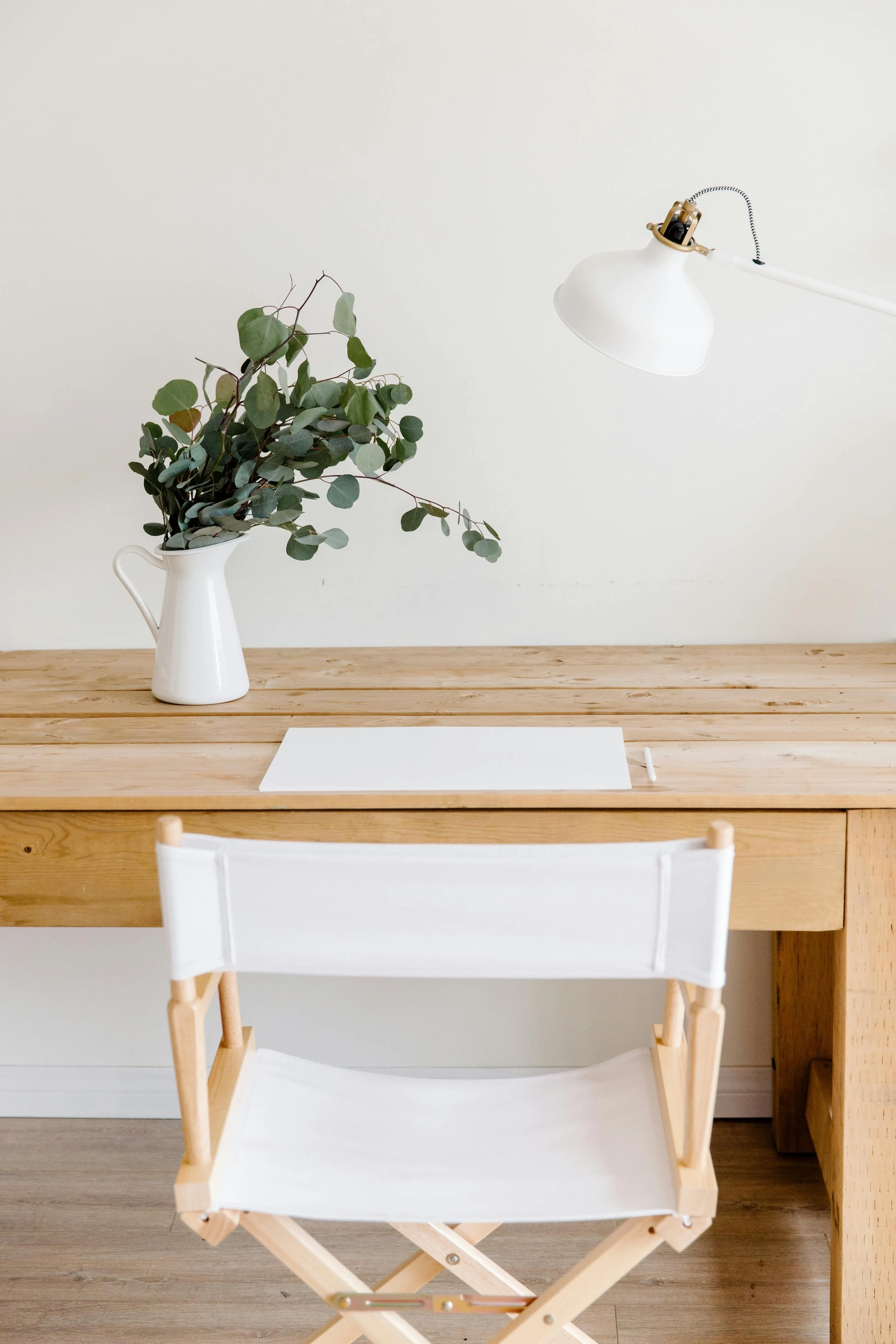A wooden desk with a white paper sheet and a white pen, a white vase with green foliage, and a white adjustable desk lamp over a plain white wall.