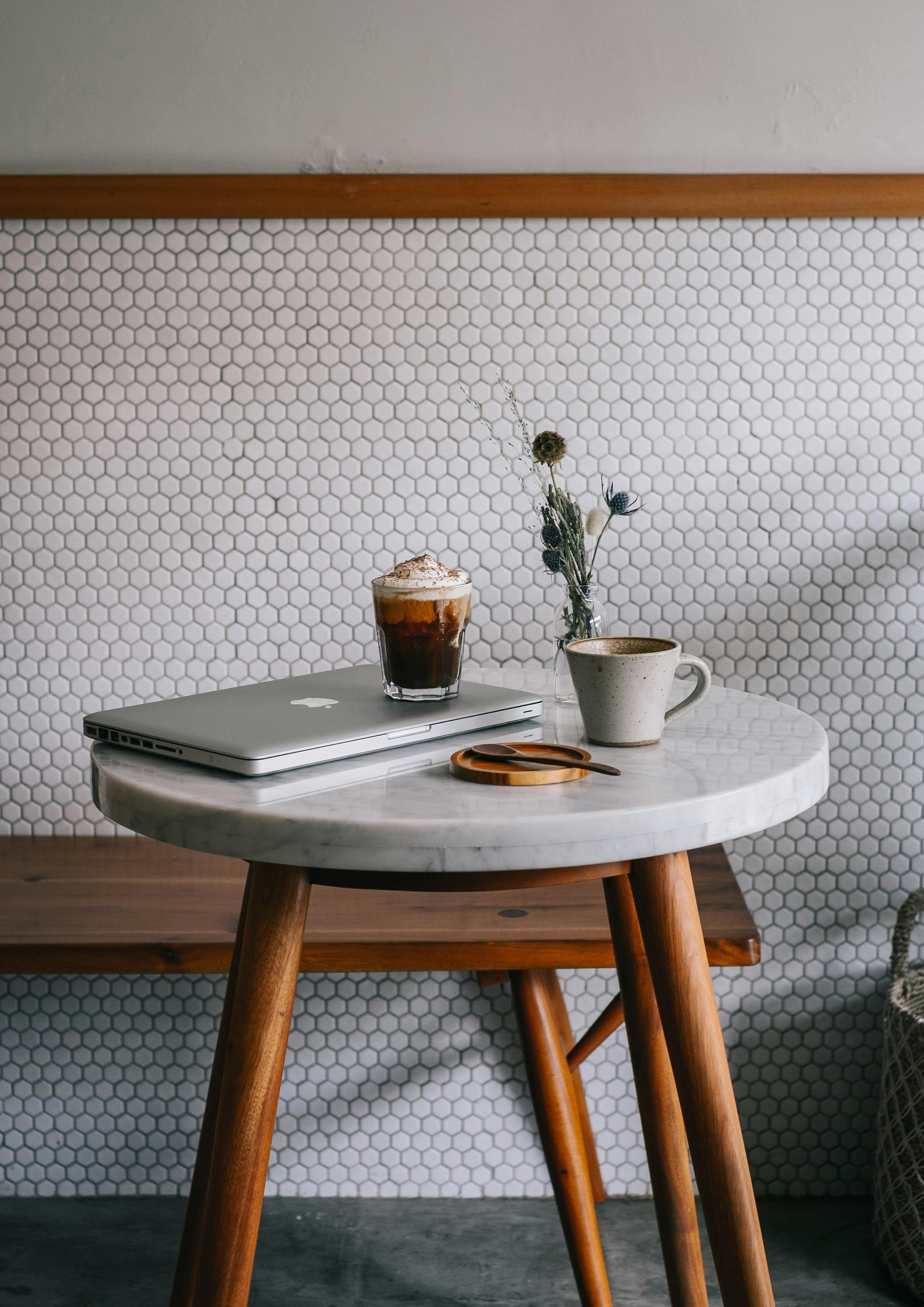 A small round marble table with a laptop, a glass of iced coffee, a ceramic mug, a wooden spoon, and a small vase with dried flowers. Behind the table is a wooden bench, and the background features a white hexagonal tile wall.