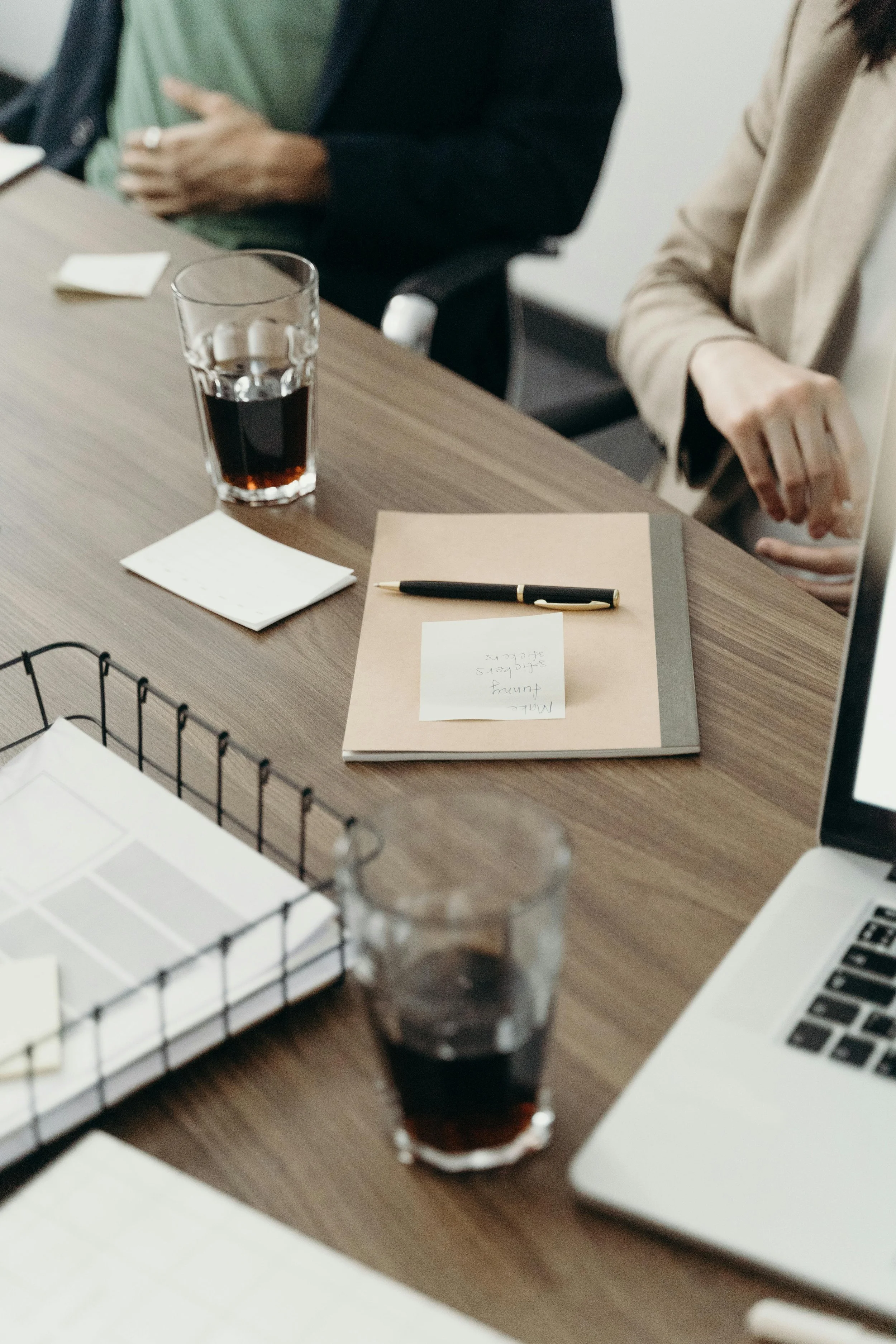 A desk with two glasses of dark soda, a notebook with a pen on top, and a laptop, with people seated nearby.