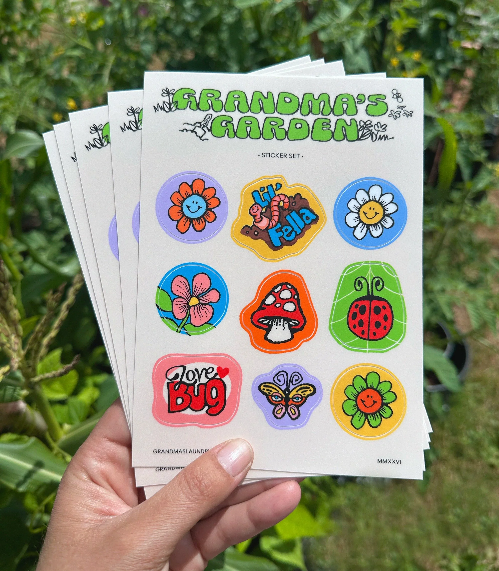 A hand holds up a stack of Grandma's Garden Sticker Sheets in front of a green planty background. The sticker sheet has 9 stickers on it: three daisies in different colors, a worm that says "Lil' Fella", a mushroom, a ladybug, a butterfly, and one th