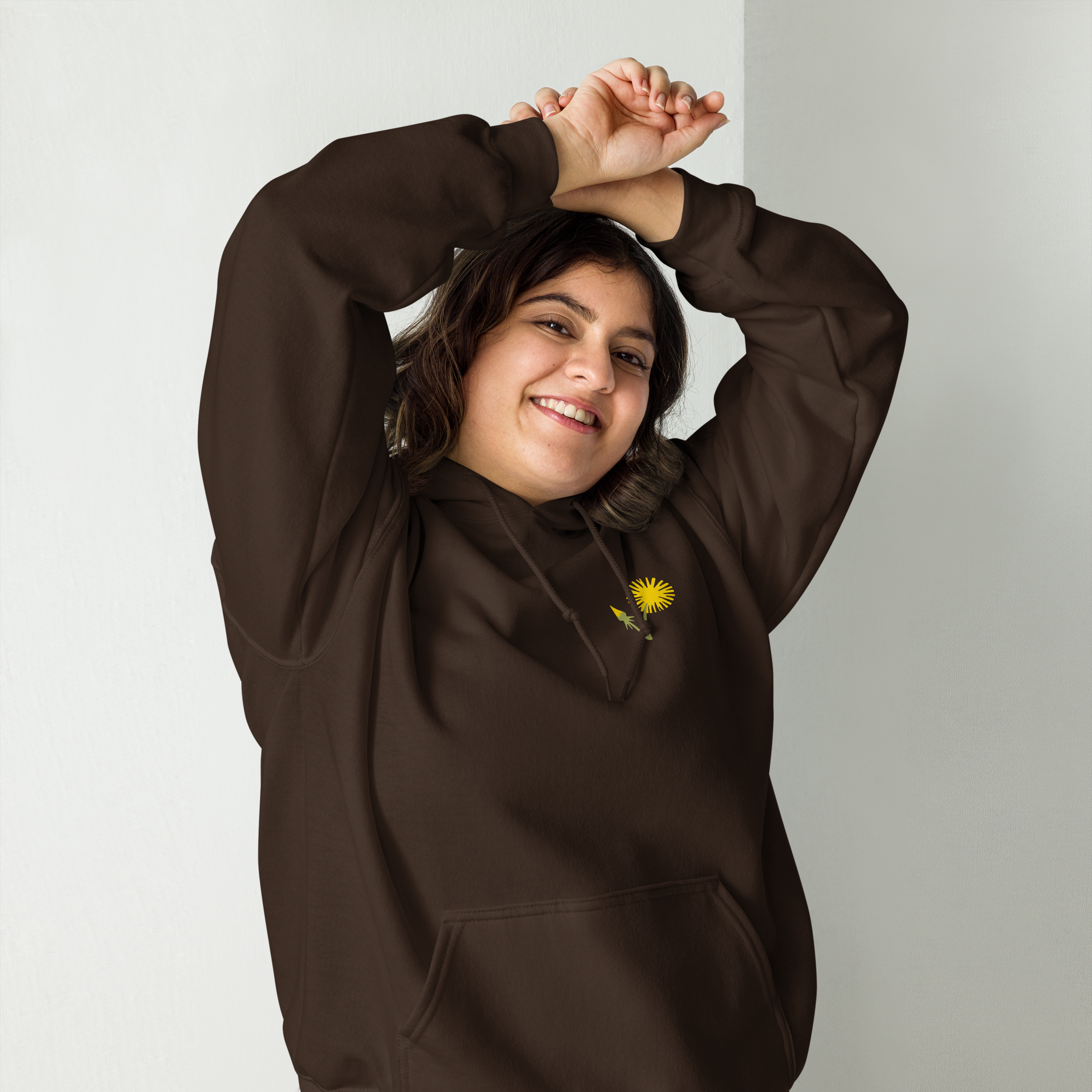 Young woman with dark hair smiling and stretching her arms above her head, wearing a brown hoodie with a yellow flower design.