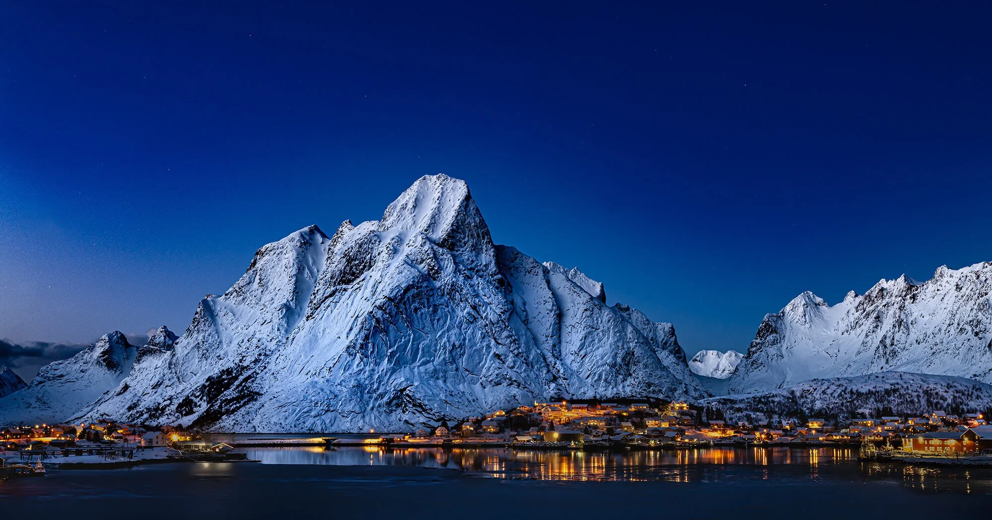 Snow-covered mountains at night with a village and reflection on a body of water in the foreground.