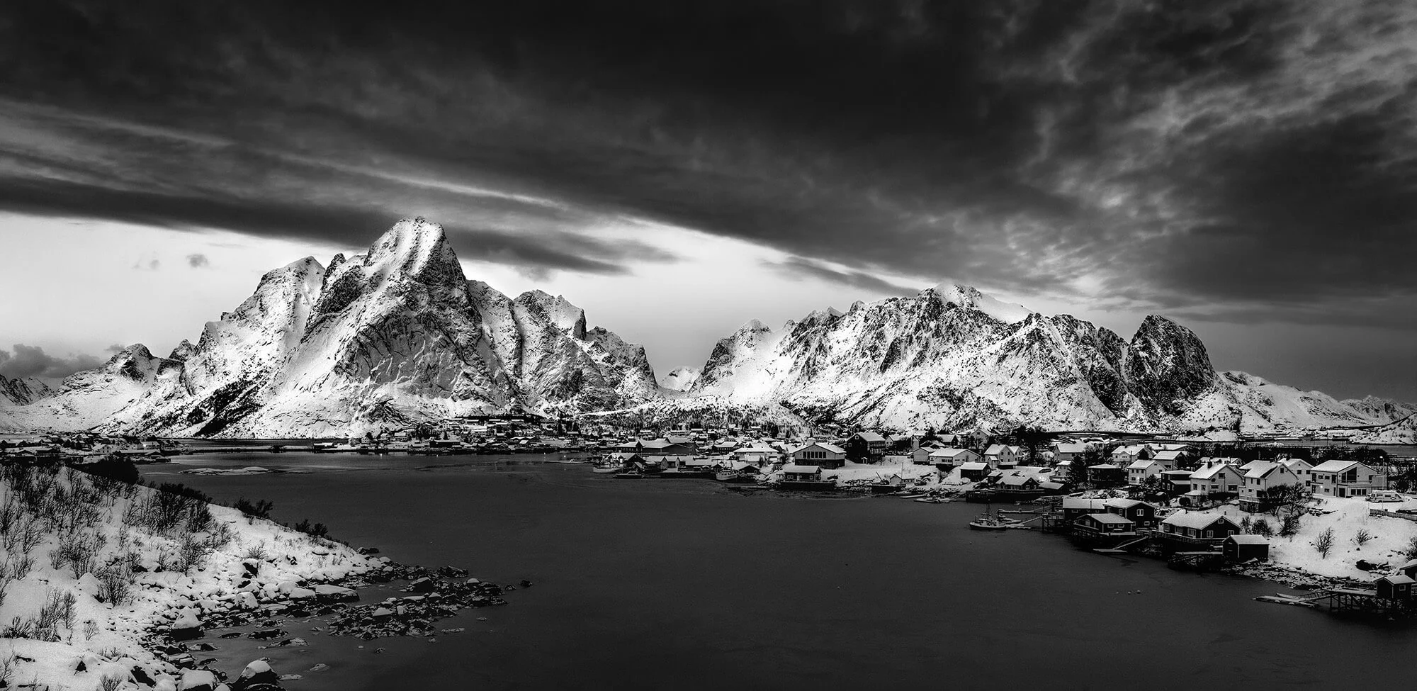 Snow-covered mountains and village in a black and white landscape with dark, cloudy sky.