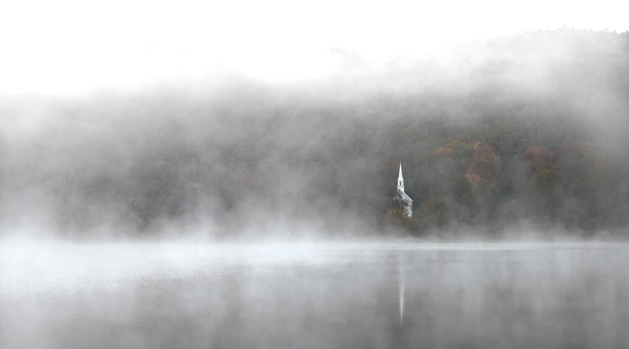A foggy lake with a small white church with a tall steeple on the opposite shore. The trees around the church are in autumn colors, partially obscured by mist.