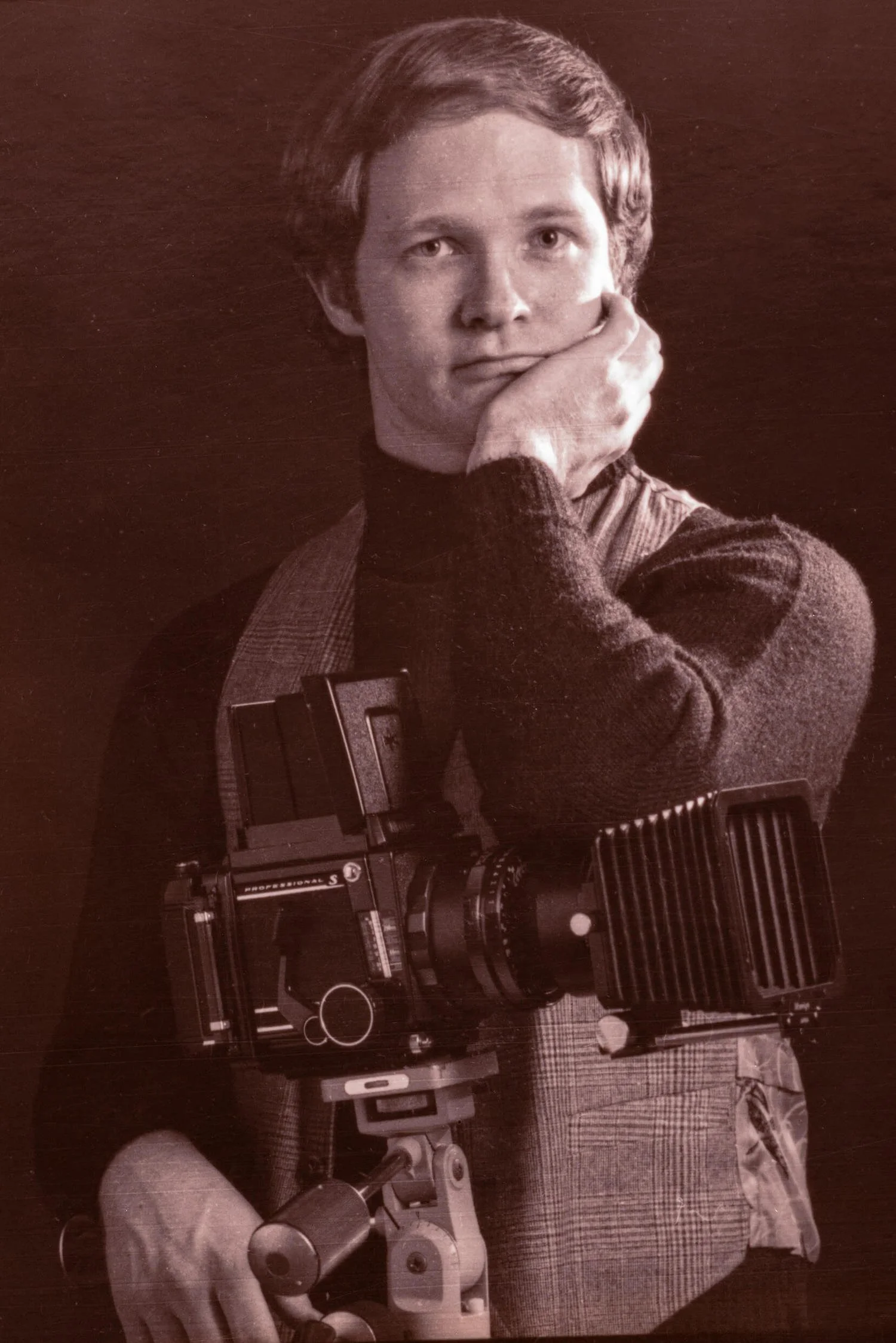 Young Steve Parham is wearing a turtleneck and standing next to a large camera on a tripod.