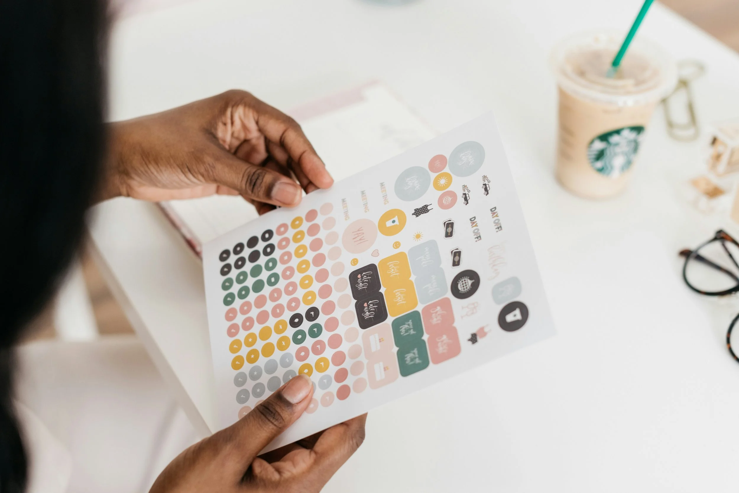 Person holding a colorful sticker sheet with various icons and labels on a white table with a Starbucks iced coffee and glasses nearby.