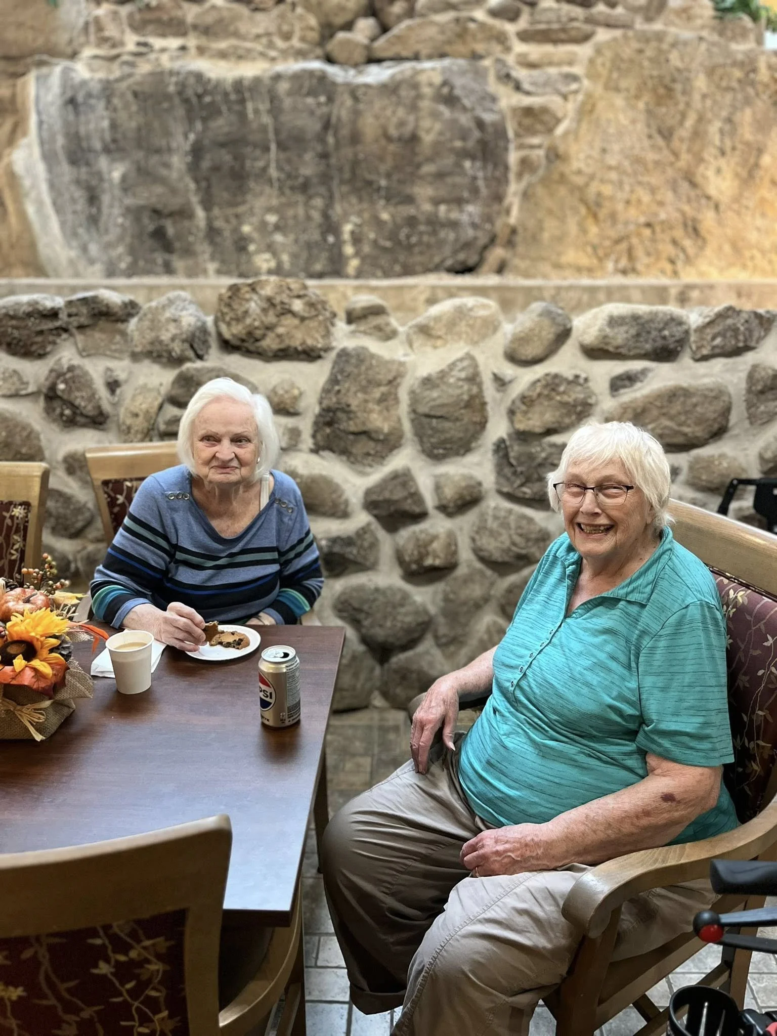 Two elderly women sitting at a dining table in a room with a stone wall background. One woman is smiling, wearing a turquoise shirt. The other woman, with gray hair, is eating a piece of cake, wearing a striped blue and black shirt. There are drinks and a floral centerpiece on the table.