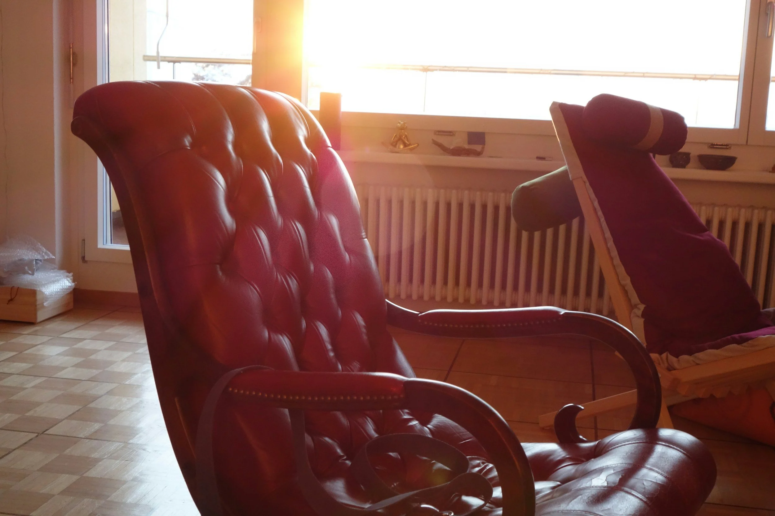Red leather armchair and purple fabric lounge chair in a sunlit room with a large window and wooden flooring.