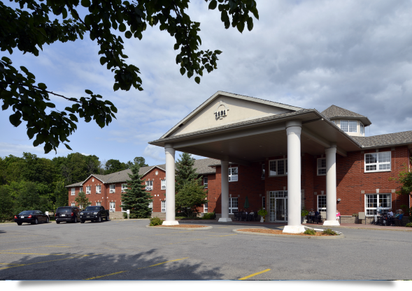 The entrance of a brick apartment complex with a large porch supported by white columns, surrounded by parking lot and trees.