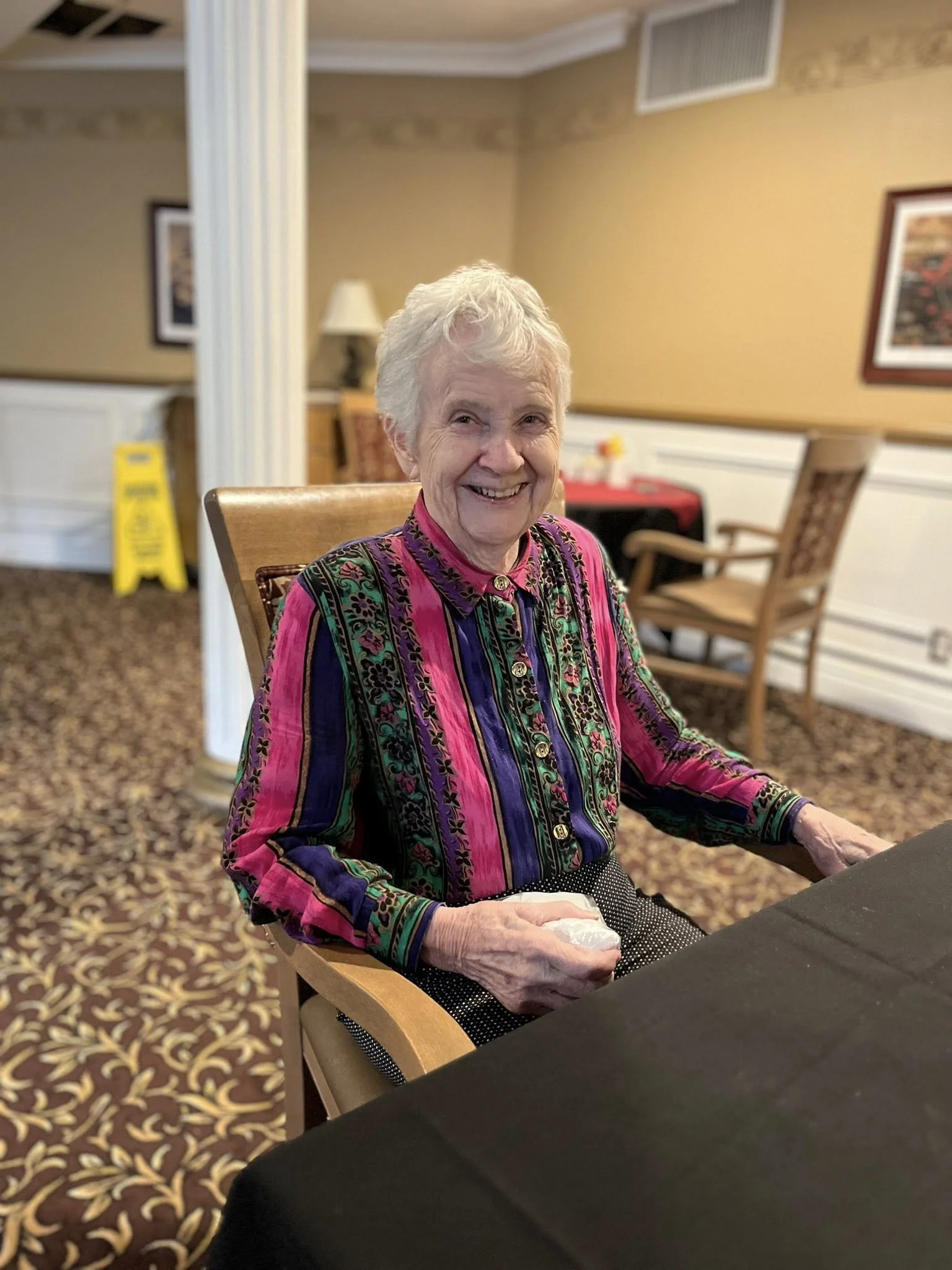 An elderly woman with white hair smiling at the camera, sitting in a chair at a dining table in a decorated room, wearing a colorful, patterned button-up shirt.