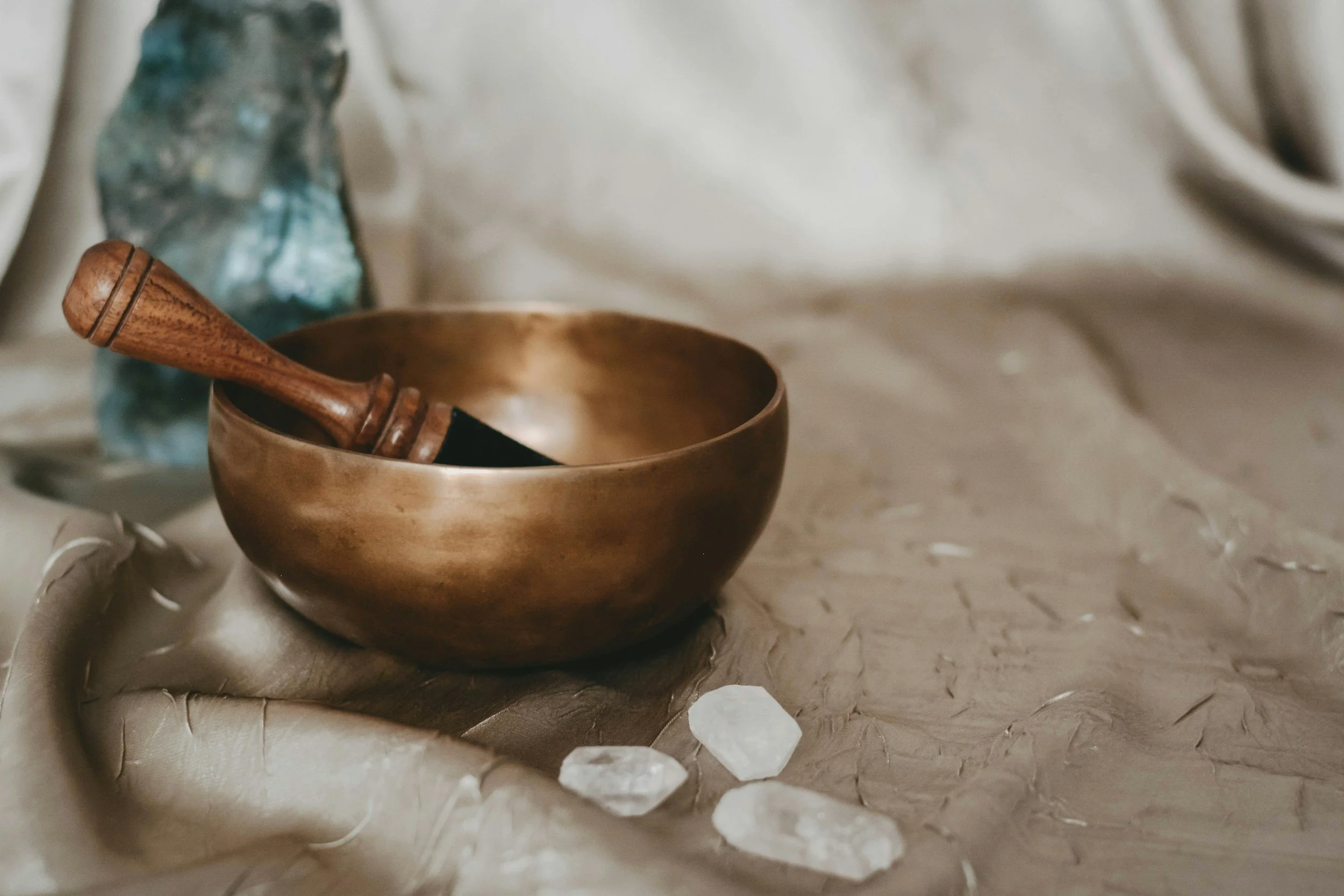 A bronze singing bowl with a wooden mallet inside, resting on crumpled beige fabric with three white crystals nearby, and a blurred blue object in the background.