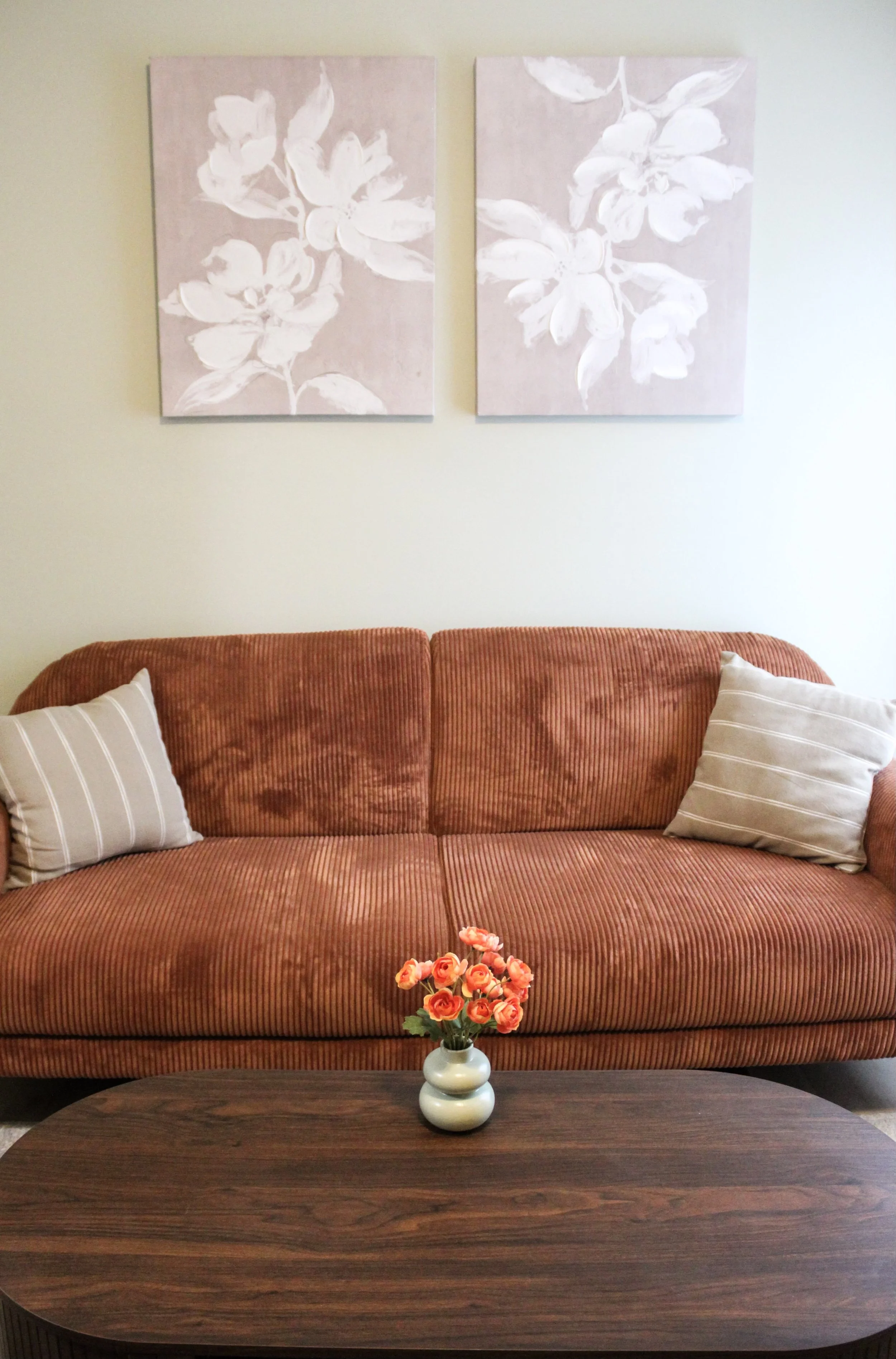 Living room with a rust-colored sofa, two beige striped pillows, a wooden coffee table with a small vase of coral-colored flowers, and two floral wall art paintings above the sofa.
