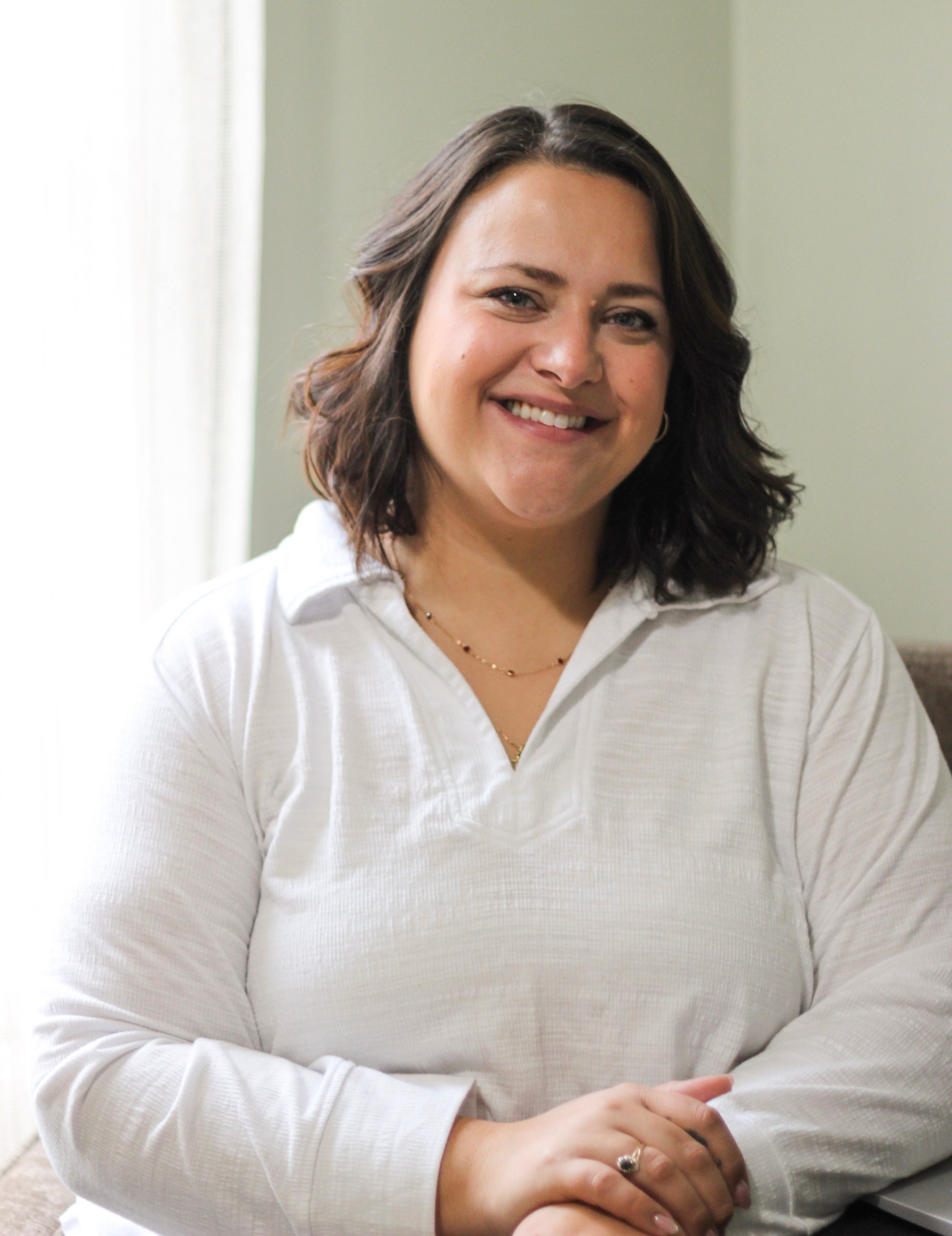 A woman with shoulder-length dark wavy hair, wearing a white long-sleeve top, smiling and sitting with hands folded on a table in a well-lit room.