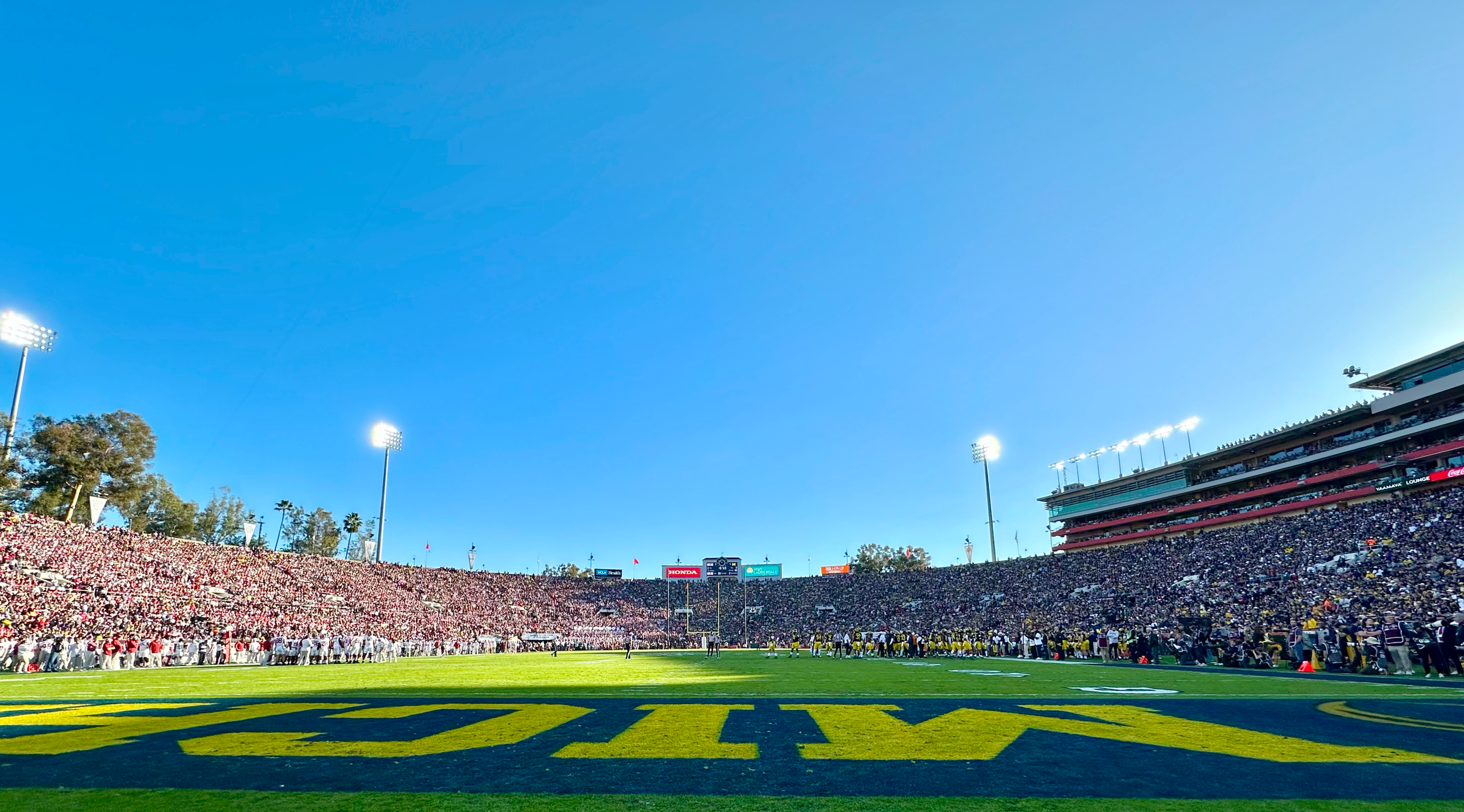 A packed football stadium with players on the field and fans in the stands, under a clear blue sky, bright stadium lights.