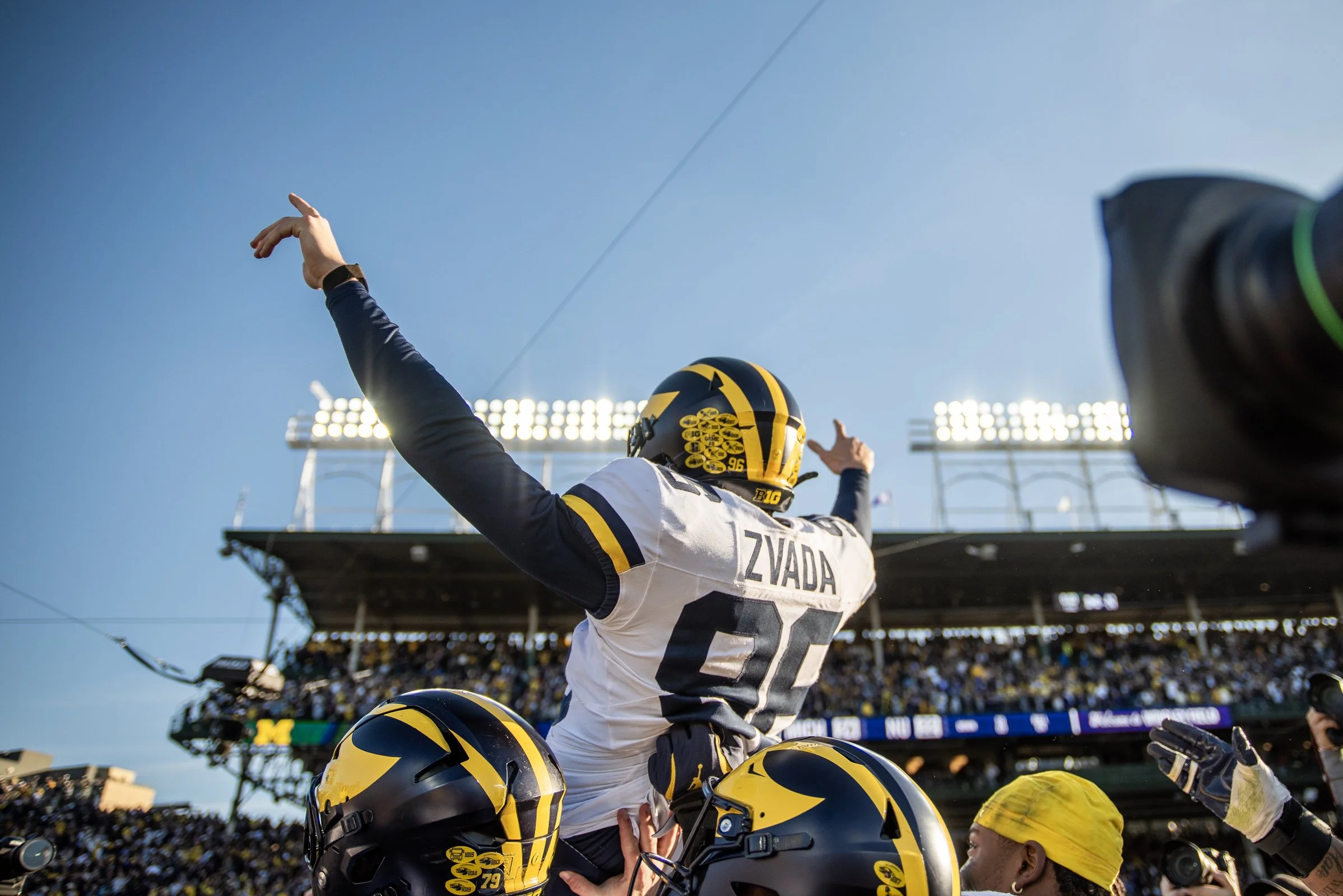 University of Michigan football player Zvizada celebrating with teammates on the field, wearing helmets and a Michigan stadium in the background under bright stadium lights.
