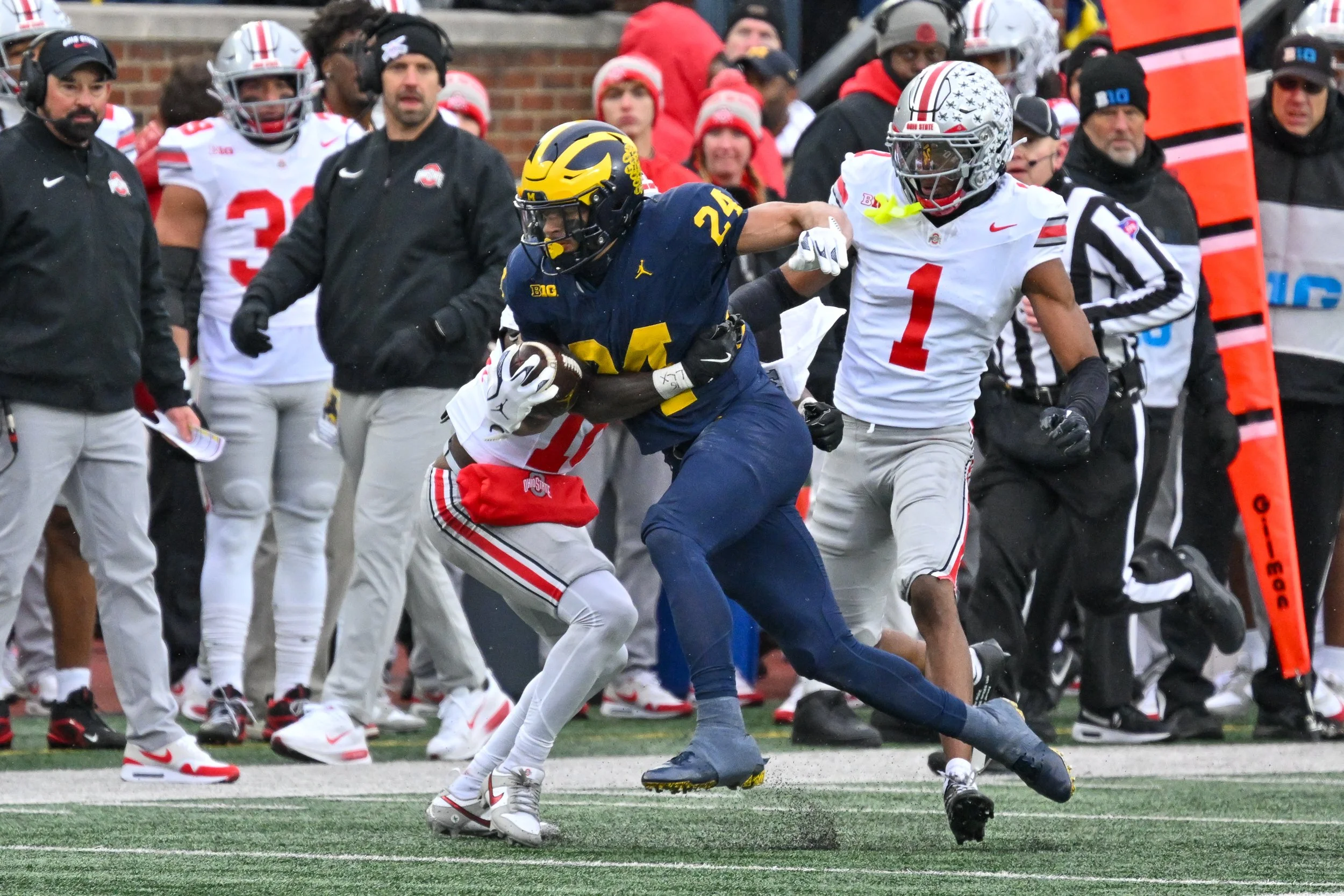 An Ohio State football player in white uniform with number 1 is being tackled by a Michigan football player in navy uniform with number 24 during a game, with multiple coaches and spectators on the sidelines.