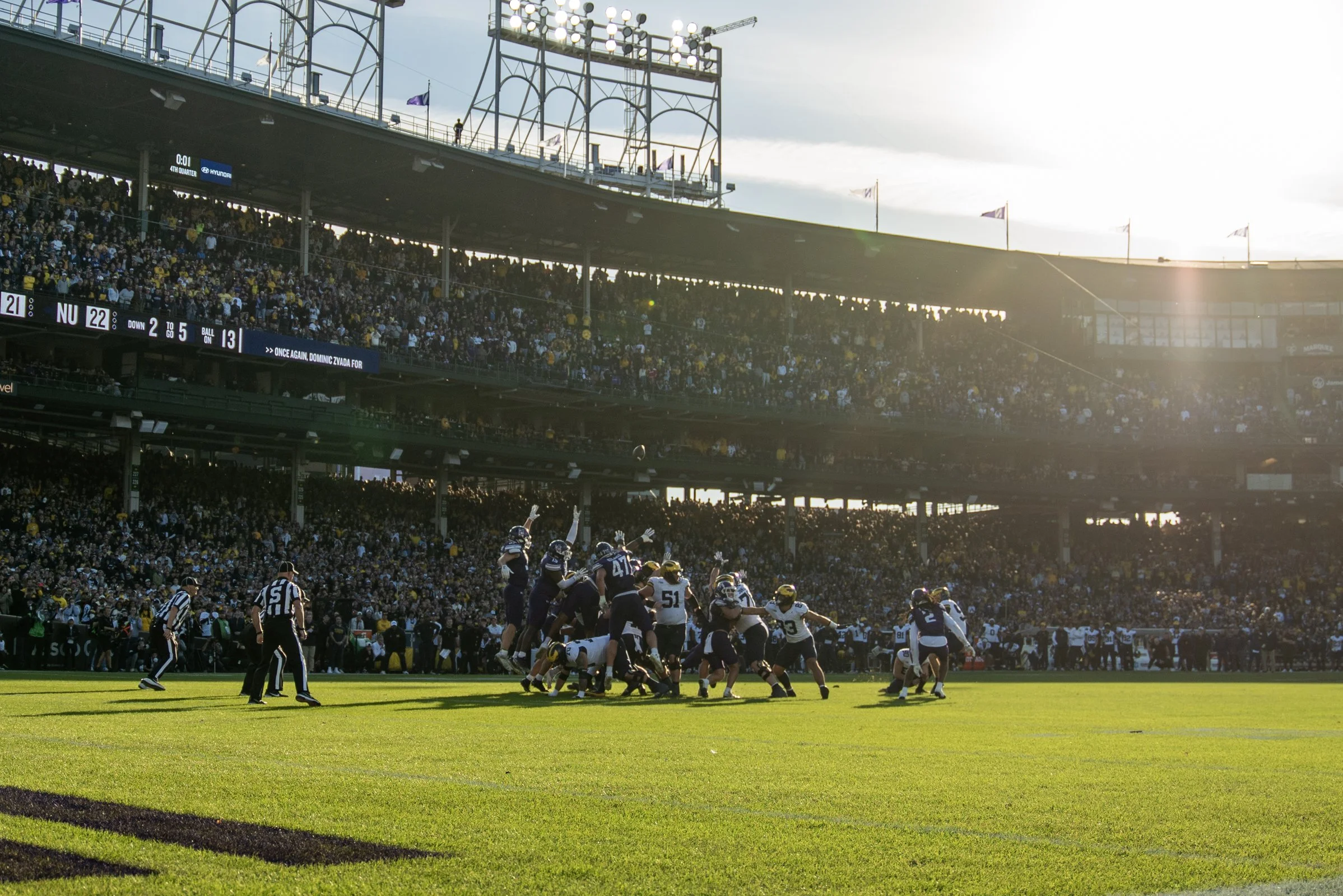A football game is in progress on a field in a large stadium filled with a crowd. Players are engaged in a play, with some on the ground and others standing, while the referee watches. The stadium is bright with sunlight, shining from the right side of the image.