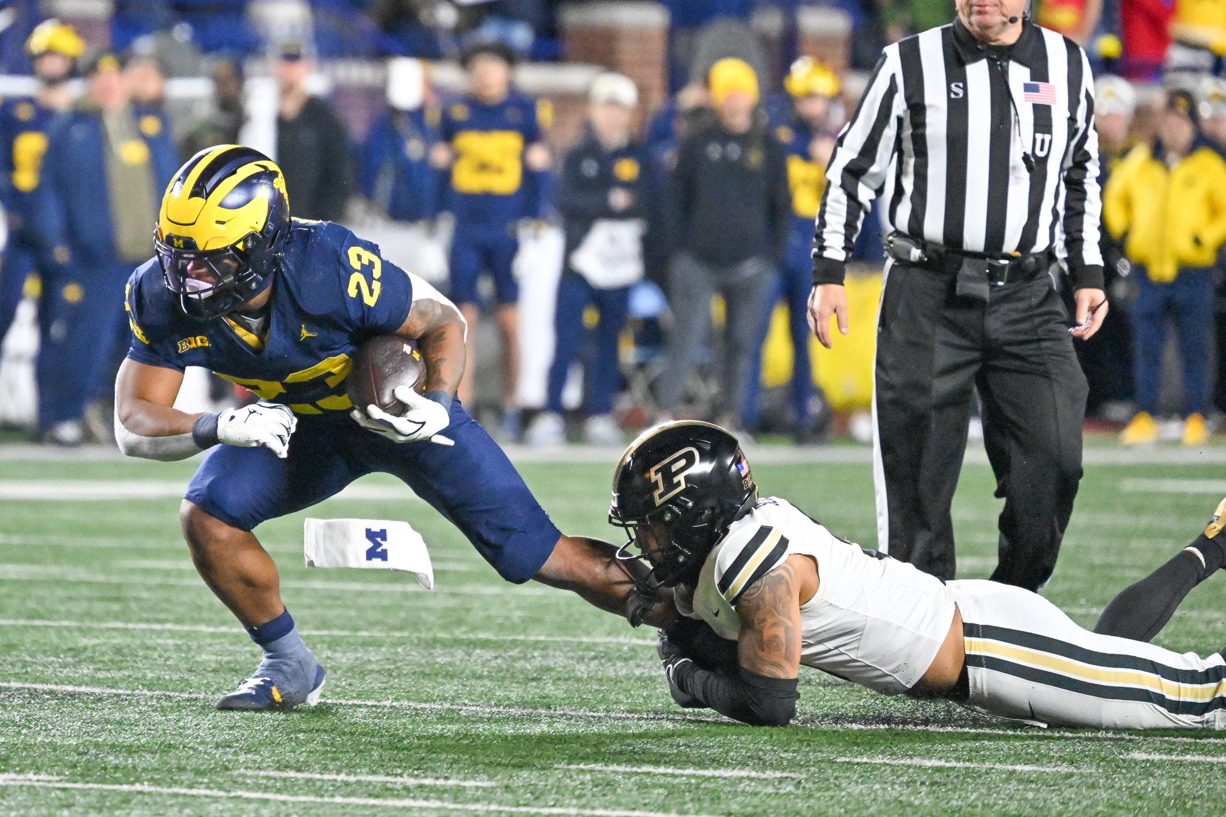 A football player from the University of Michigan wearing a blue uniform with the number 23, running with the ball while being tackled by a player from Purdue wearing a white uniform. An official in a black and white striped shirt is standing nearby, and there are spectators in the background.