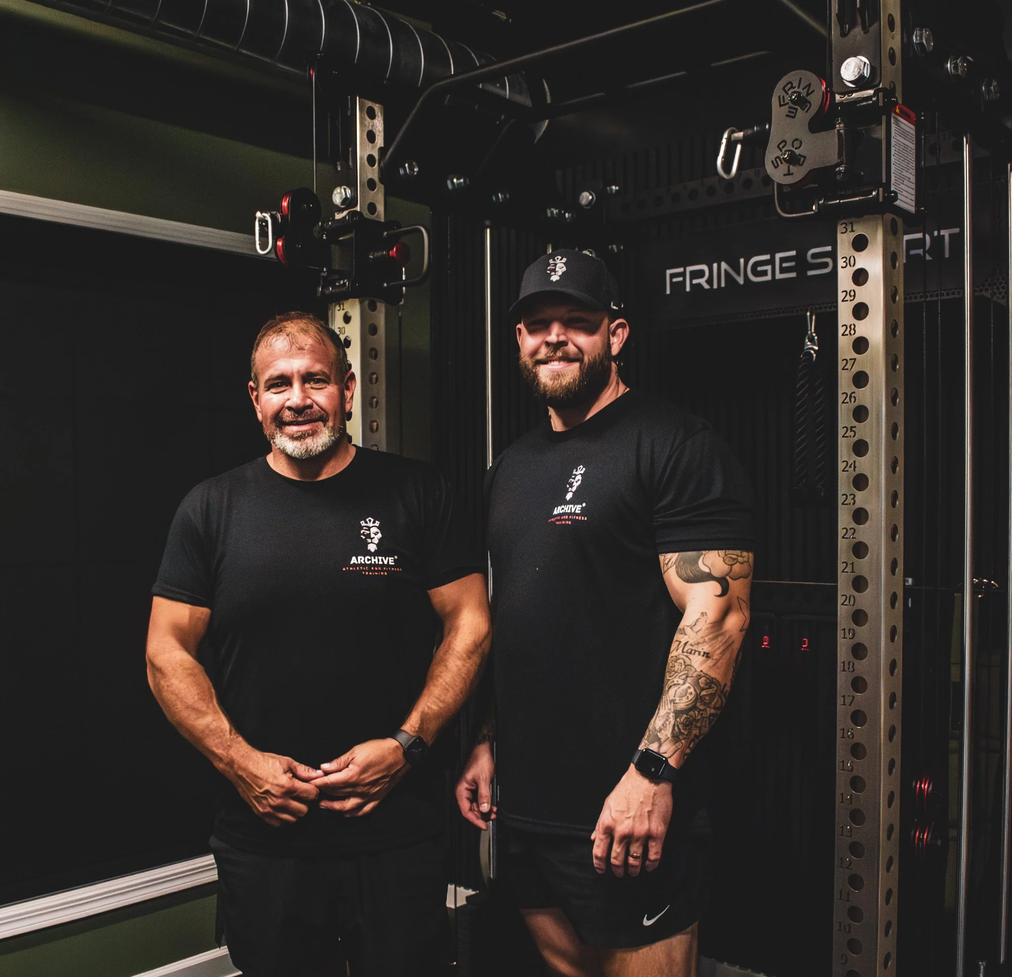 Personal trainer and client standing on the gym floor in front of strength equipment. Image highlights professional coaching staff and structured training environment.