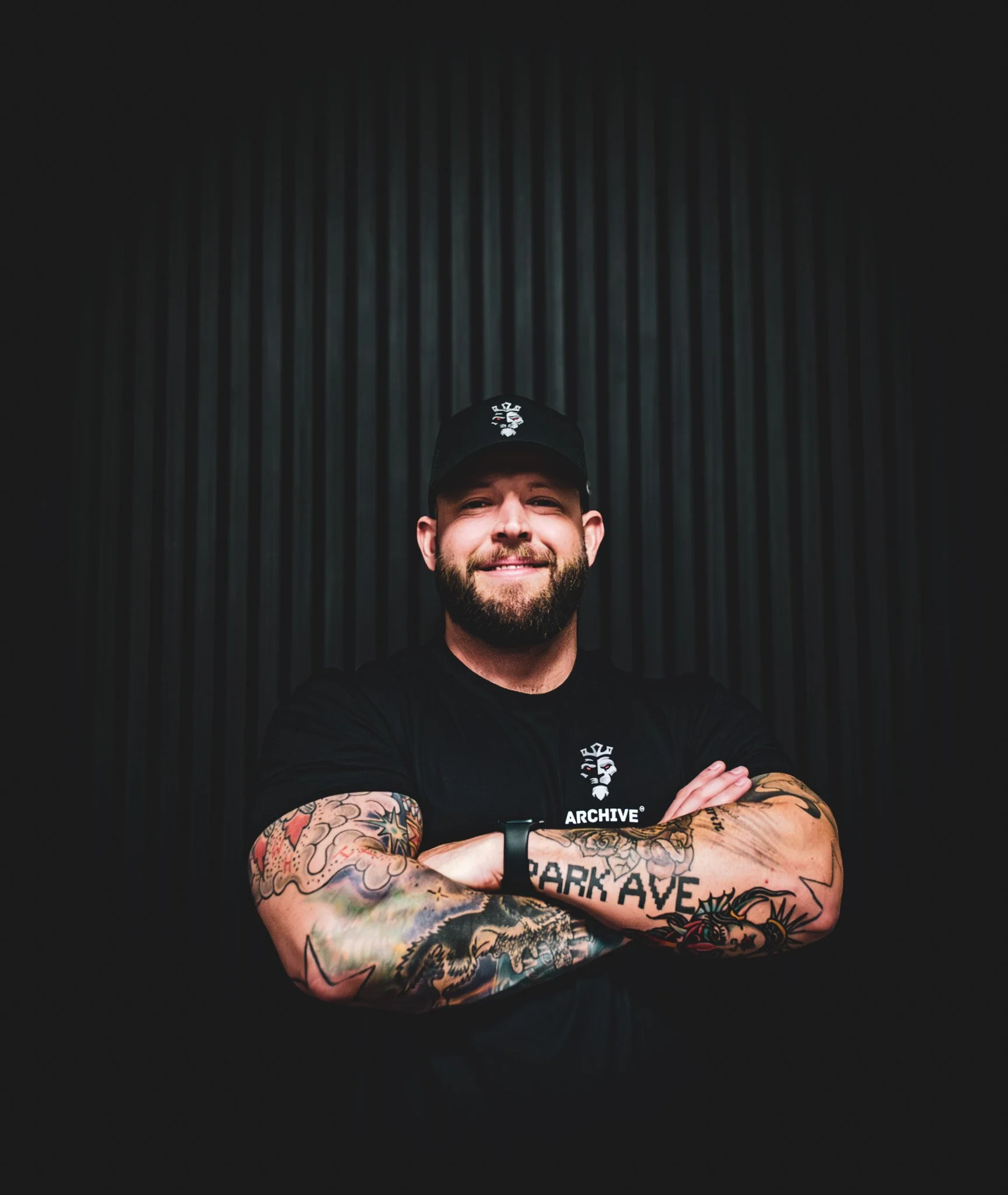 A smiling man with a beard and tattoos on his arms, wearing a black cap and black T-shirt, standing with arms crossed in front of a dark, textured background.