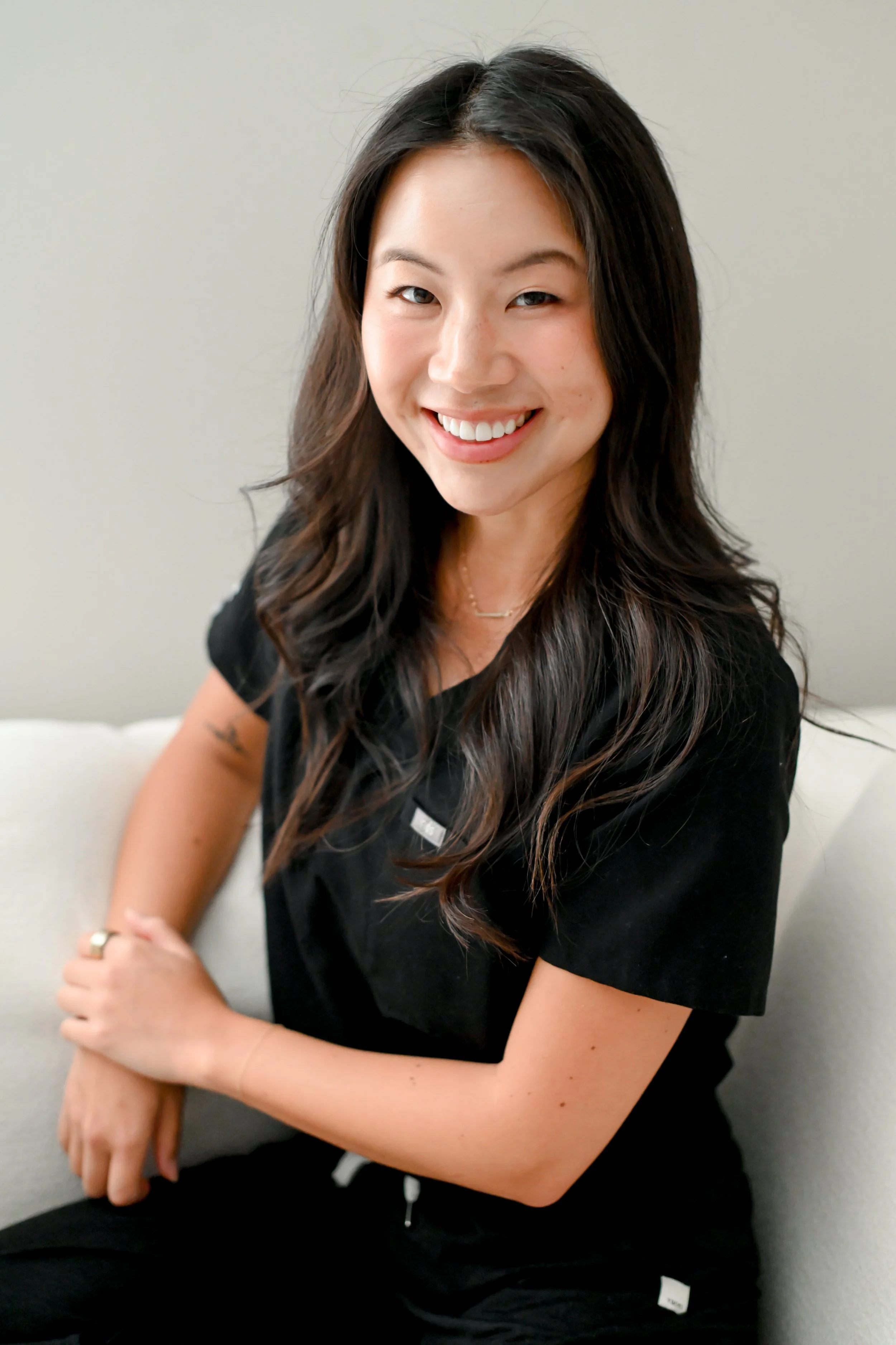 A young woman with long dark hair smiling at the camera, sitting on a white couch in a neutral-colored room, wearing a black shirt.