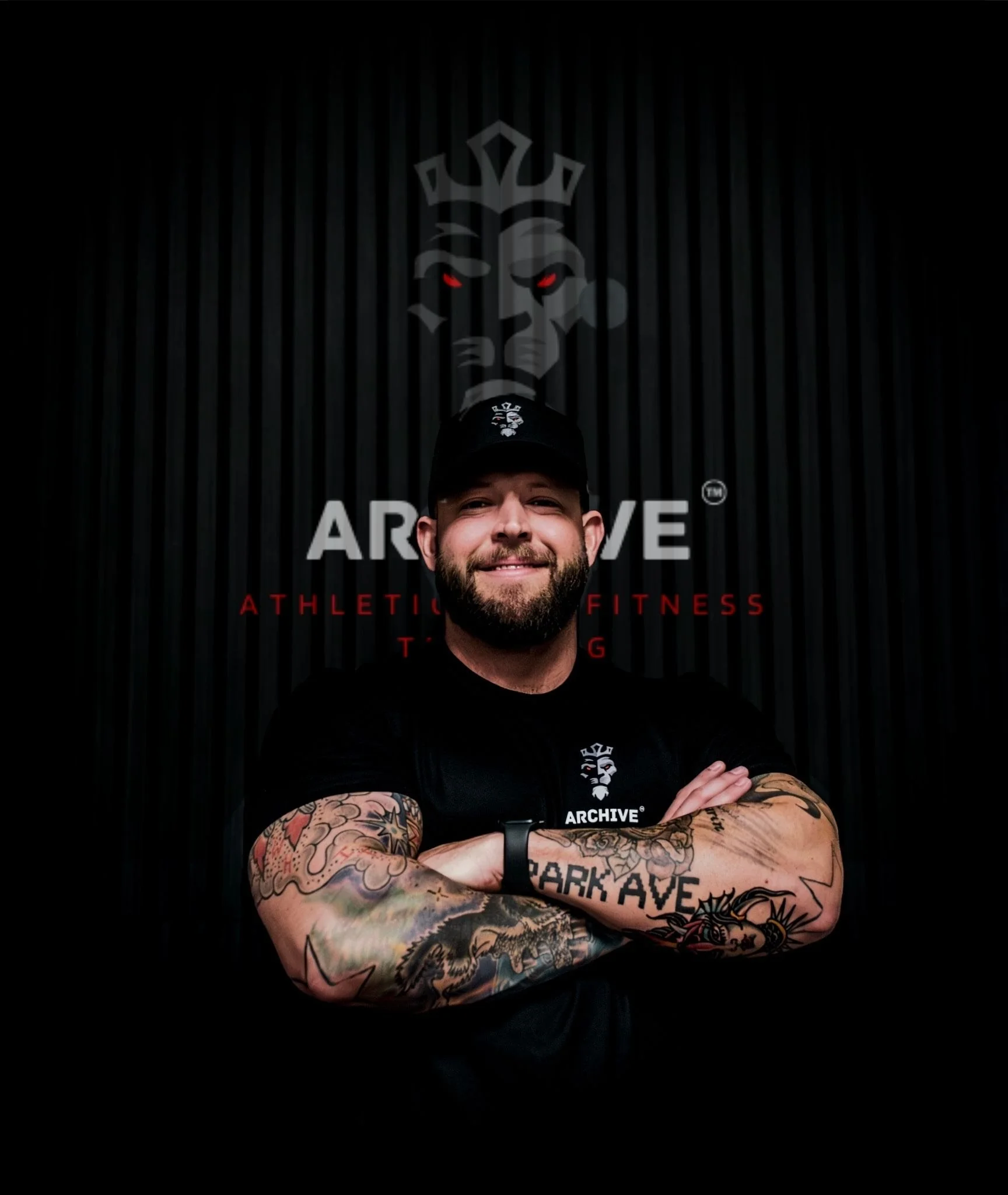 Man with a beard and tattoos wearing a black hat and black shirt, standing with arms crossed in front of a dark backdrop with a lion logo and the words 'ARCHIVE ATHLETICS FITNESS TRAINING'.